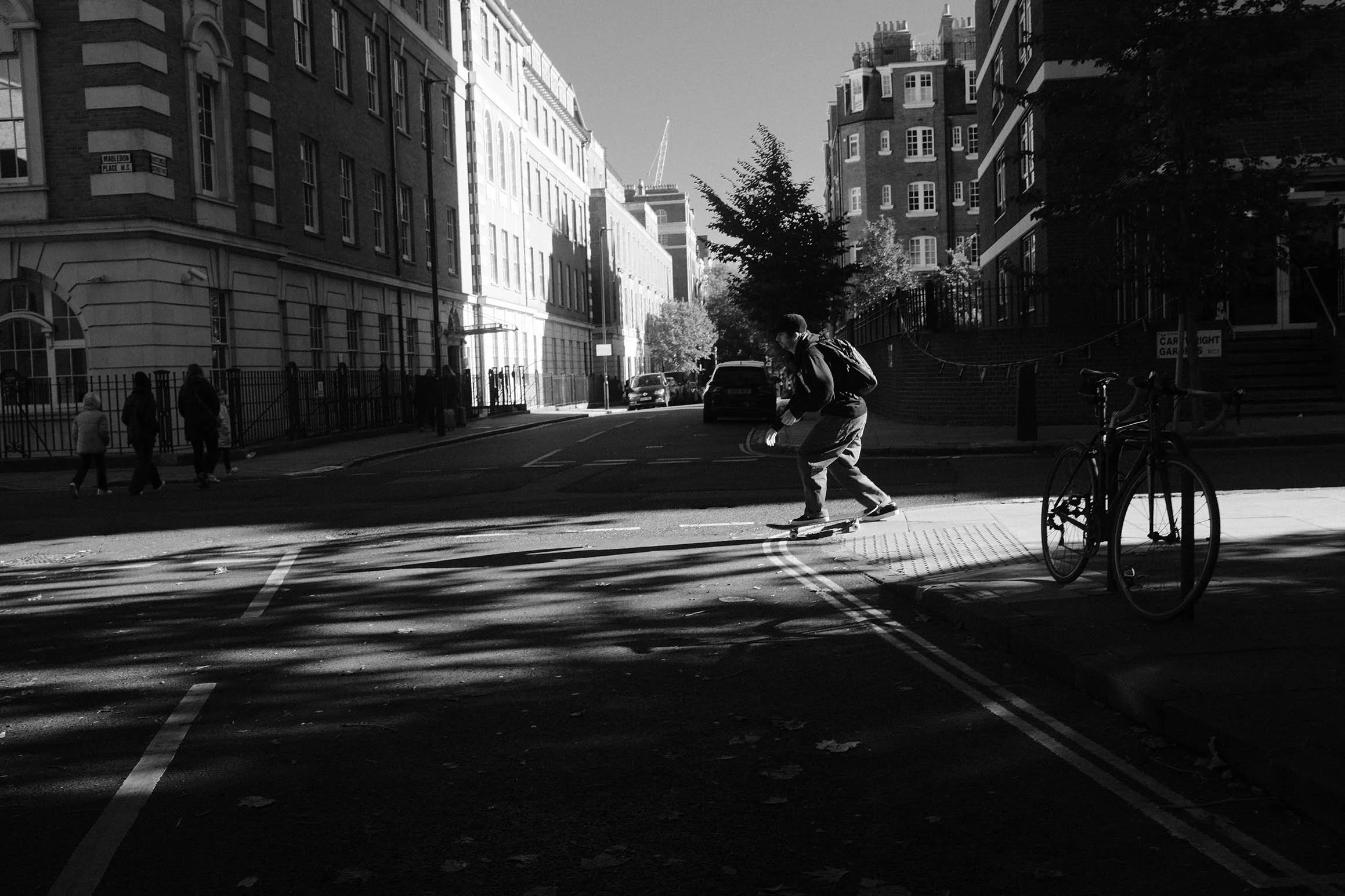 Un jeune homme fait du skateboard dans une rue citadine, entourée de bâtiments résidentiels, avec une bicyclette garée sur le trottoir à droite et des enfants marchant au fond à gauche. La photo est en noir et blanc.