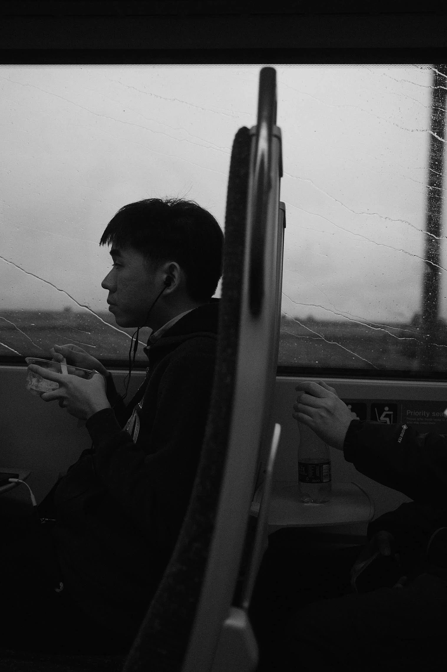 Jeune homme assis dans un bus regardant un téléphone, avec un autre passager dont la main repose sur une table, en arrière-plan une fenêtre avec vue sur un paysage urbain.