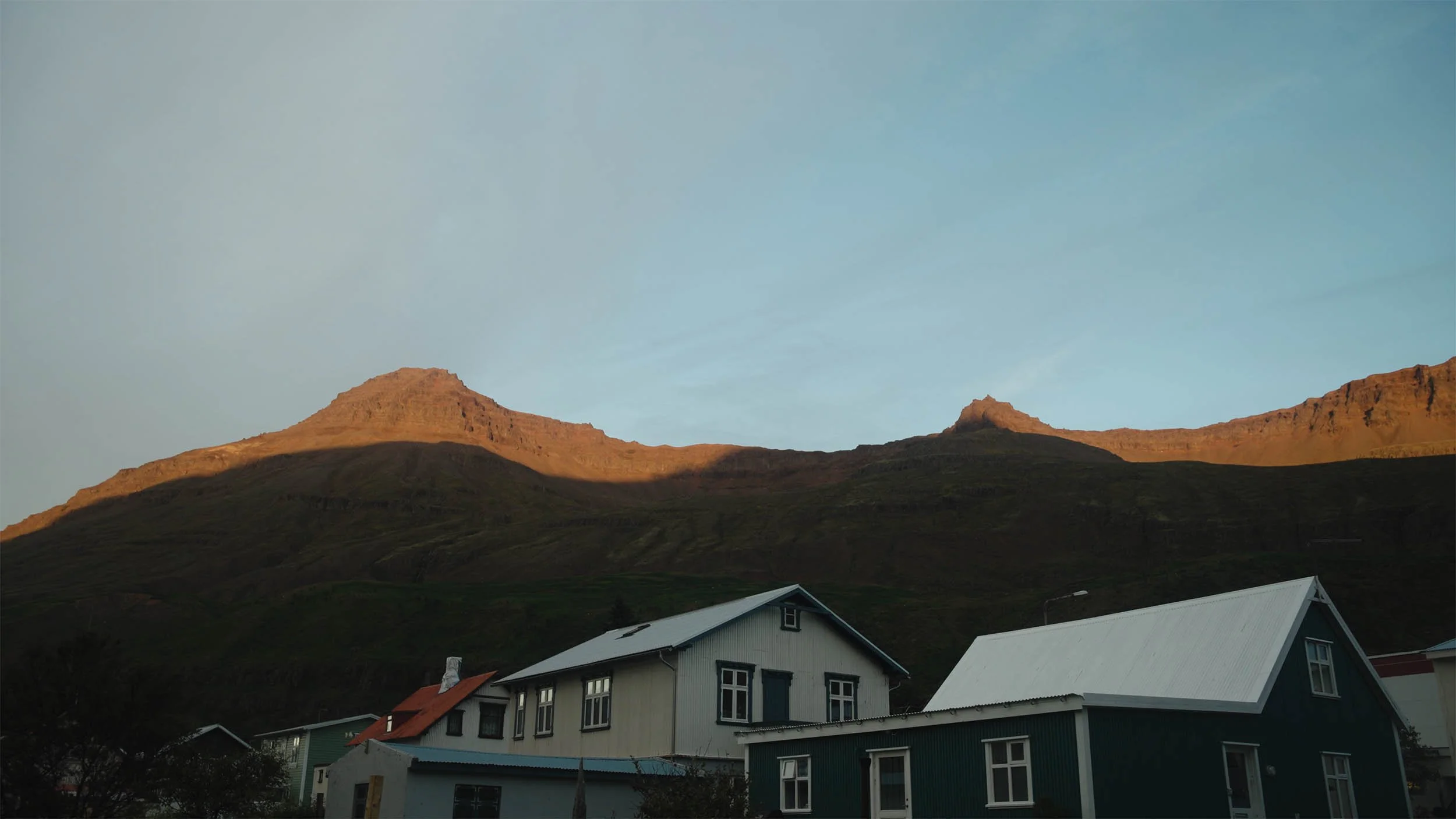 Maisons en bois à faible hauteur avec des toits inclinés, montagne en arrière-plan, ciel clair, en fin de journée ou début de soirée.