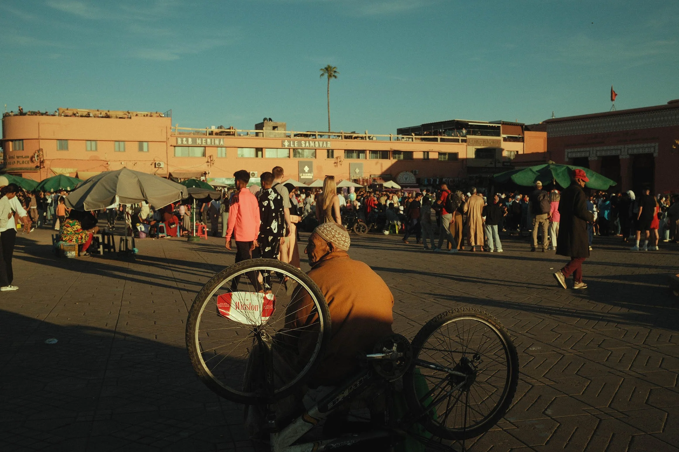 Un homme âgé en vélo sans roue assis dans une place animée, avec de nombreux vendeurs et passants, bâtiments en arrière-plan, et un ciel clair.