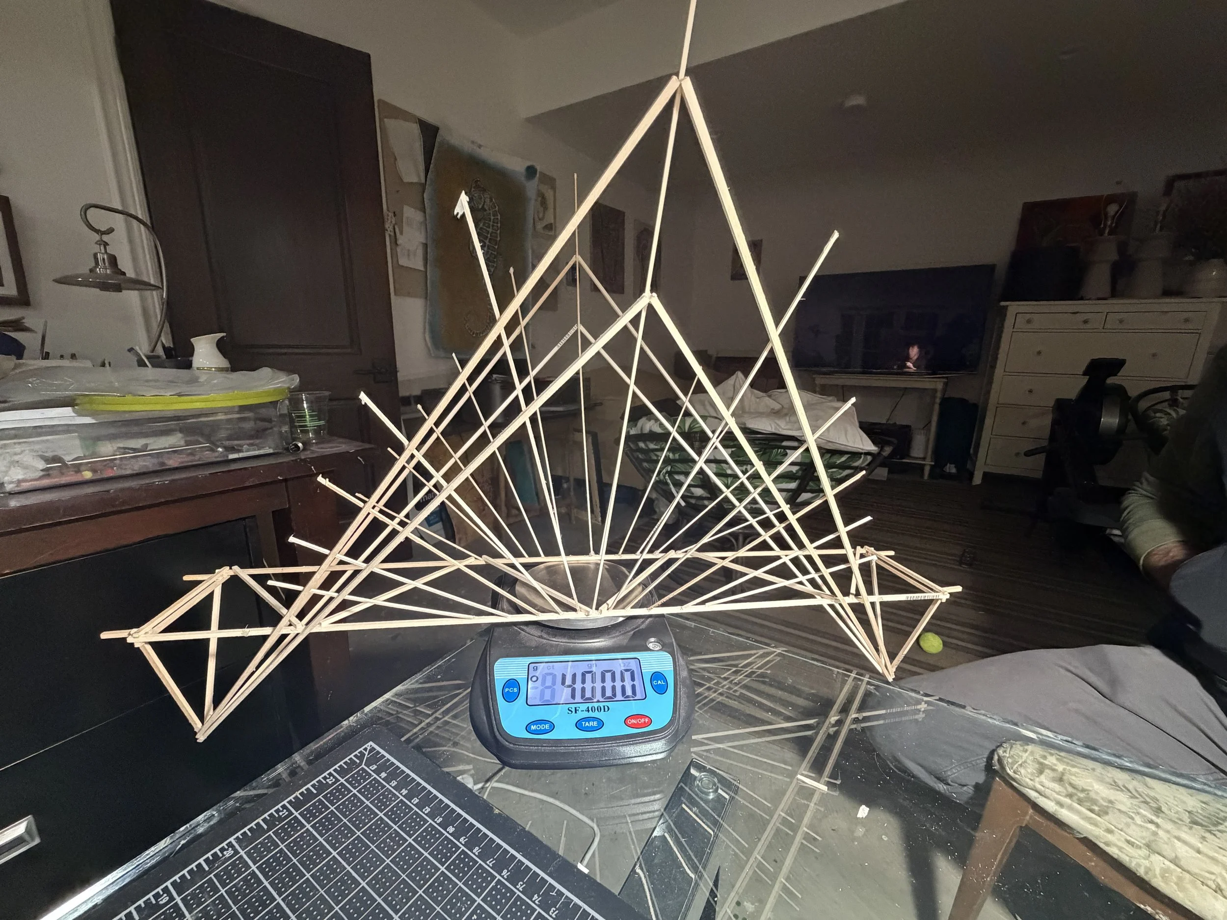 A wooden geometric sculpture or structure placed on a digital scale showing 84.00 grams in a cluttered room with furniture, a television, and various objects.