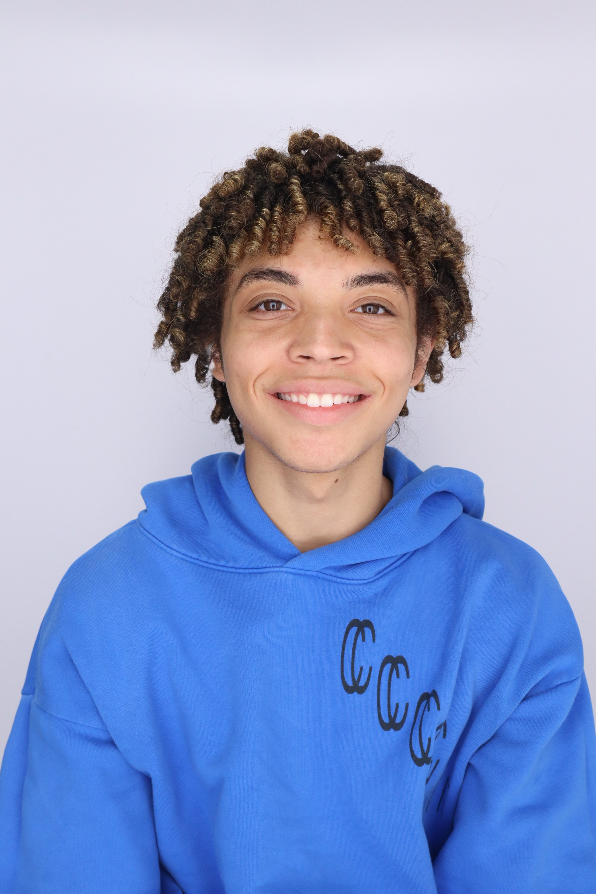 Close-up of a smiling young person with curly hair in a blue hoodie against a plain background.