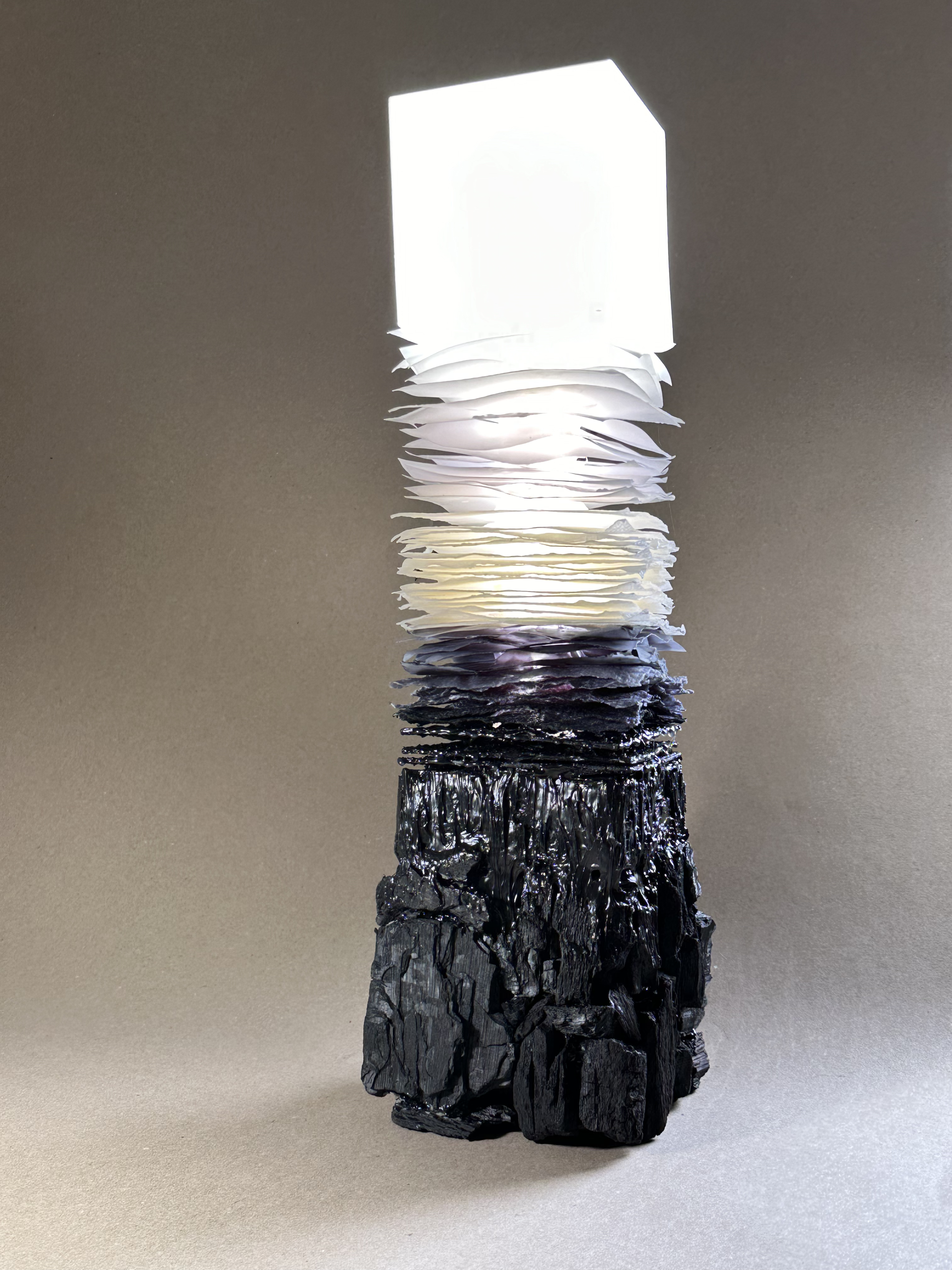 A layered sculpture with a black base, a middle section of white paper-like materials, and an upper part of a illuminated square on a gray background.