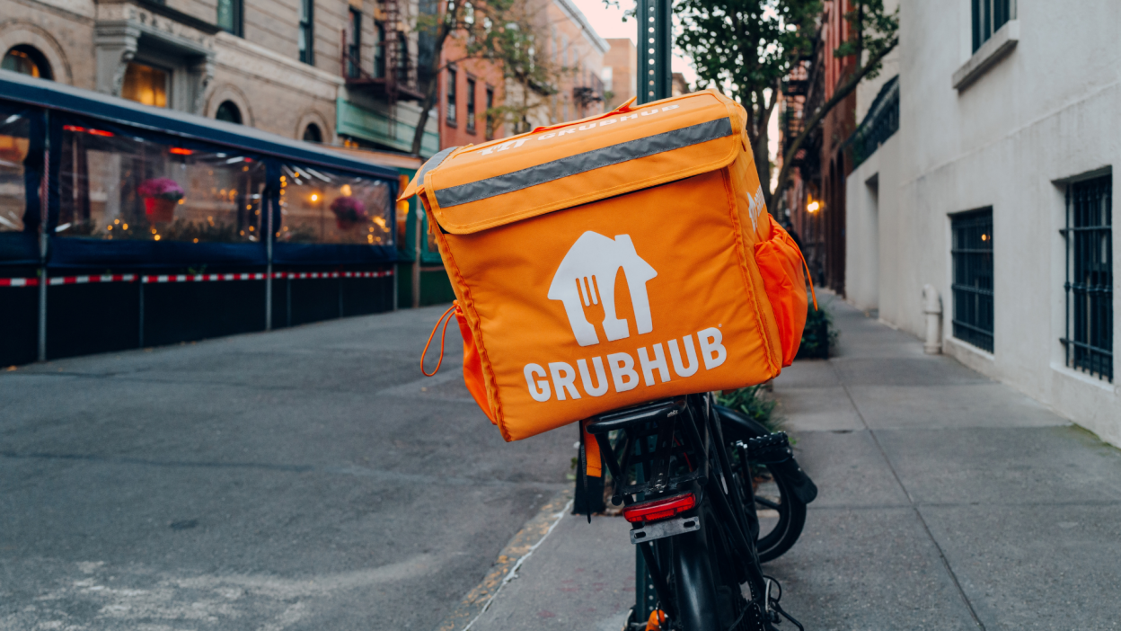 An orange Grubhub delivery bag attached to a bicycle on a city sidewalk.