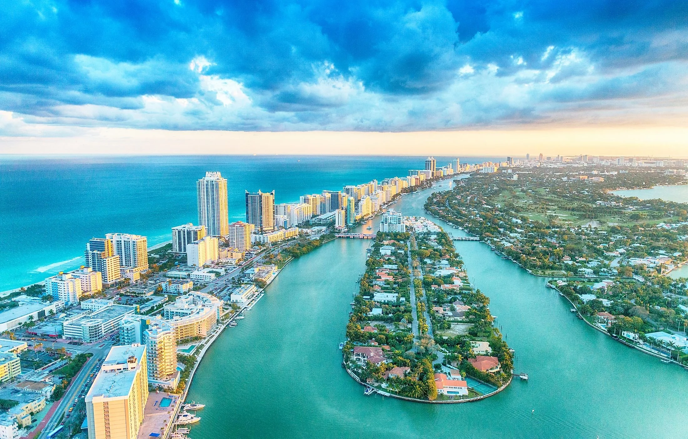 An aerial view of a cityscape with high-rise buildings along a coastline, and a winding river through residential areas, under a partly cloudy sky at sunset.