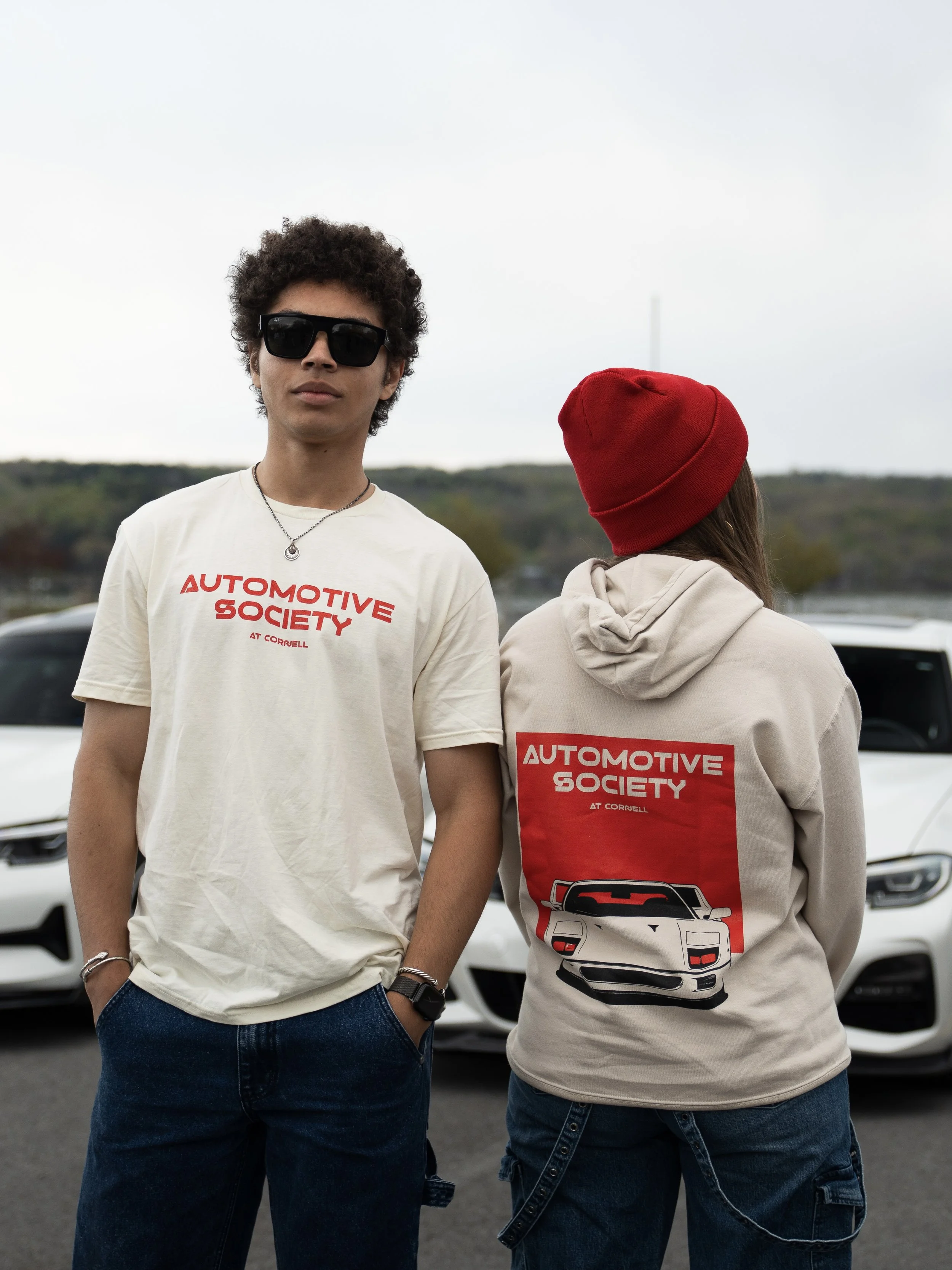 Two people standing in front of cars during daytime. One person is facing forward with sunglasses, wearing a white T-shirt with red text that reads 'Automotive Society at Cornell' and has their hands in their pockets. The other person has their back to the camera, wearing a red beanie and a cream hoodie with a red and white 'Automotive Society at Cornell' logo and a car graphic on the back. The background features a cloudy sky and a scenic landscape.