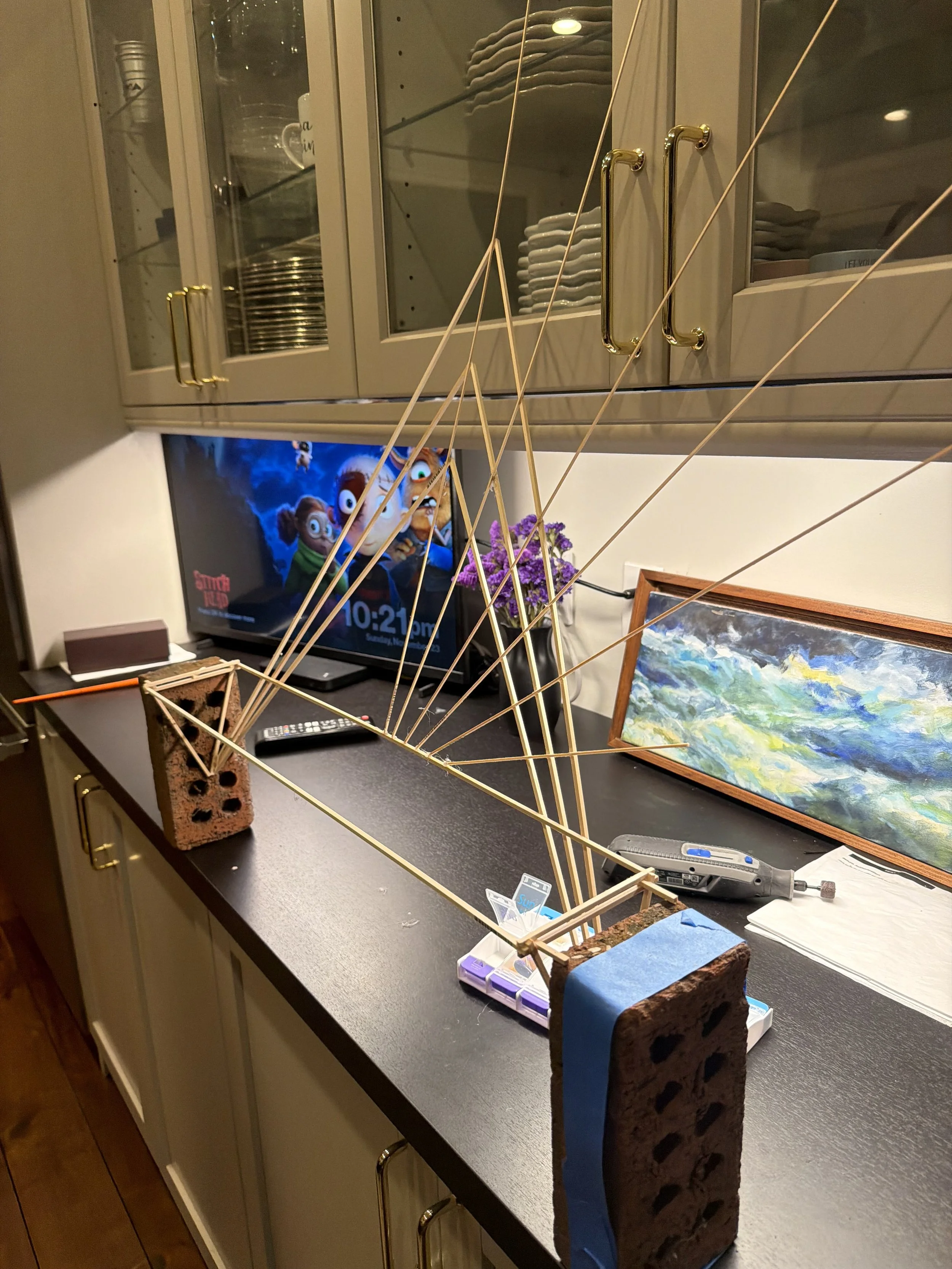A workspace with a partially built model bridge on the counter, held in place with bricks and tape. Behind it, a TV displays a cartoon show featuring animated animals. There are shelves with dishes and bowls, a framed landscape painting, and some purple flowers.
