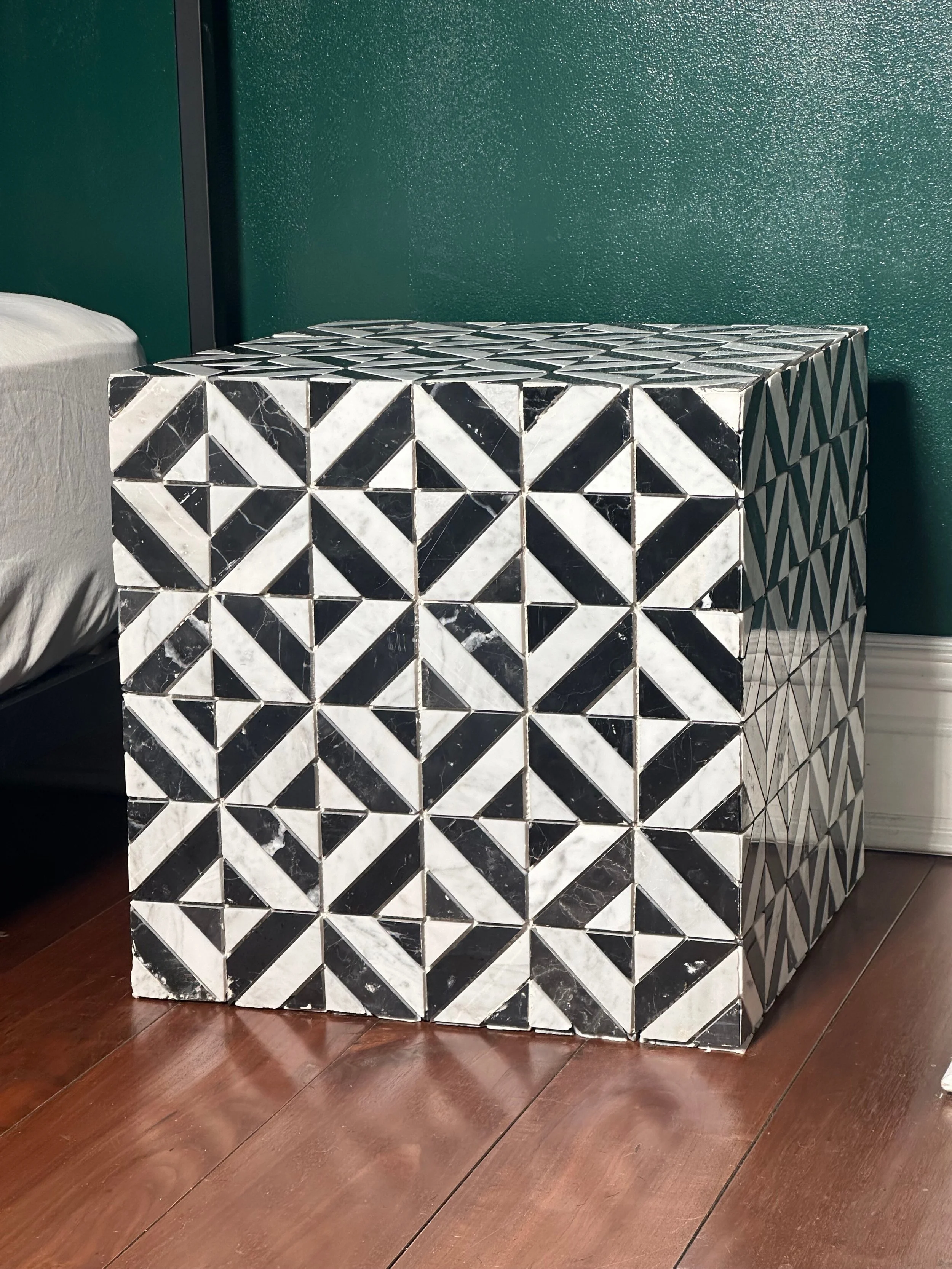 A black and white patterned tile side table next to a bed with a beige pillow, against a green wall with a textured finish, on a wooden floor.