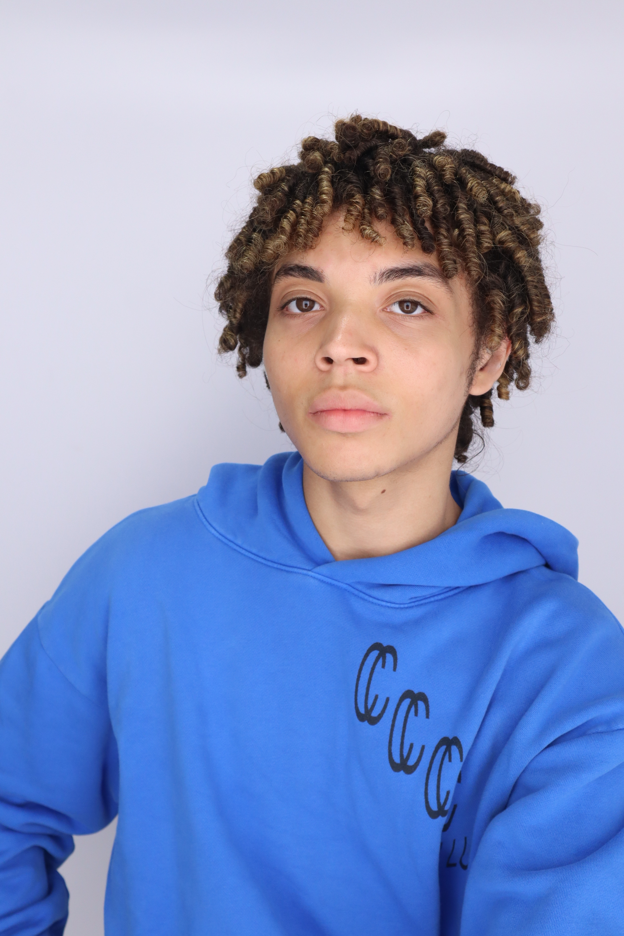 A young person with light skin and curly hair wearing a blue hoodie with black graphic design, standing against a plain white background.