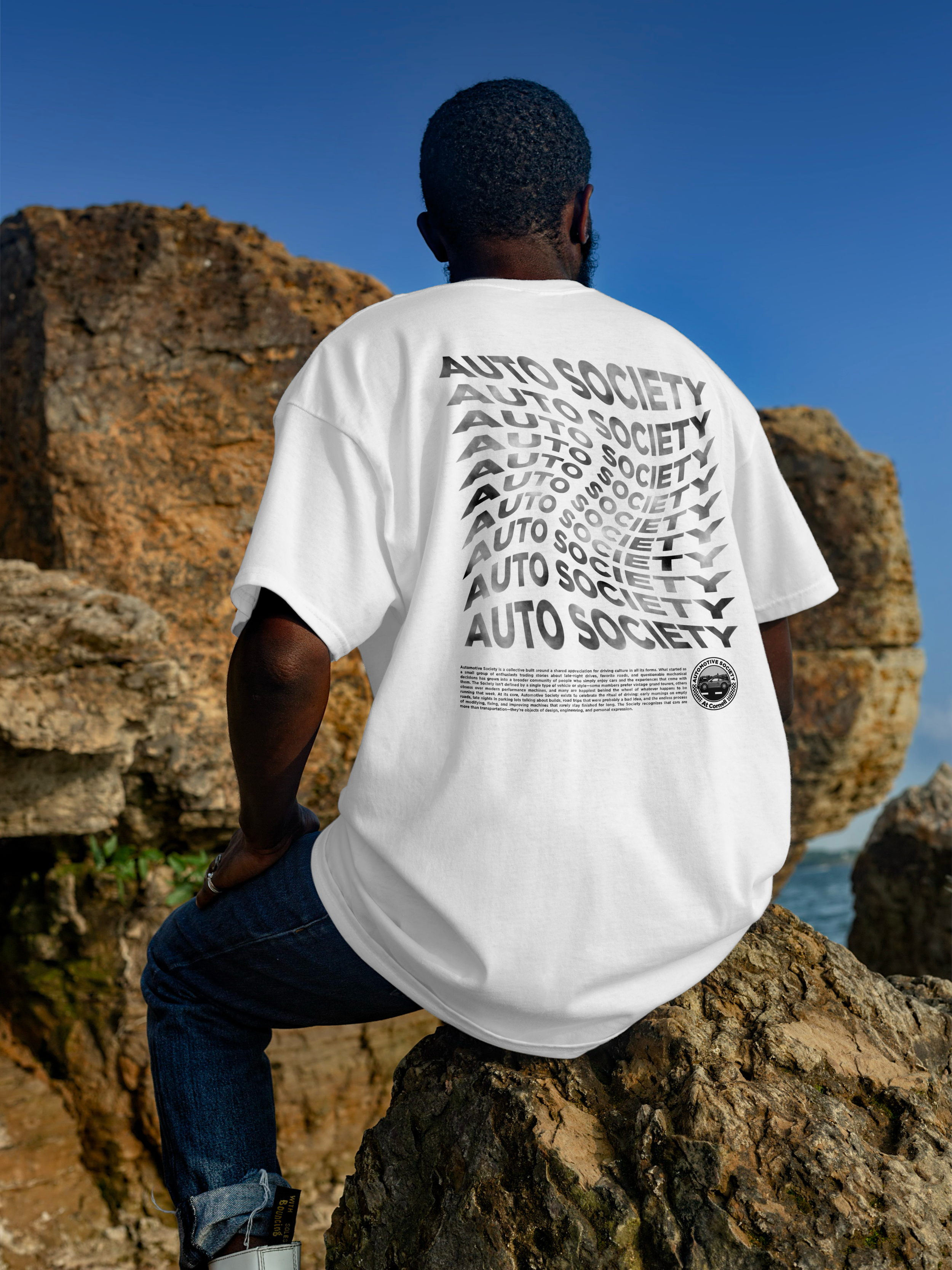 Person wearing a white T-shirt with a repeating black print that says 'AUTO SOCIETY,' sitting on a large rock outdoors with a blue sky and rocky landscape in the background.