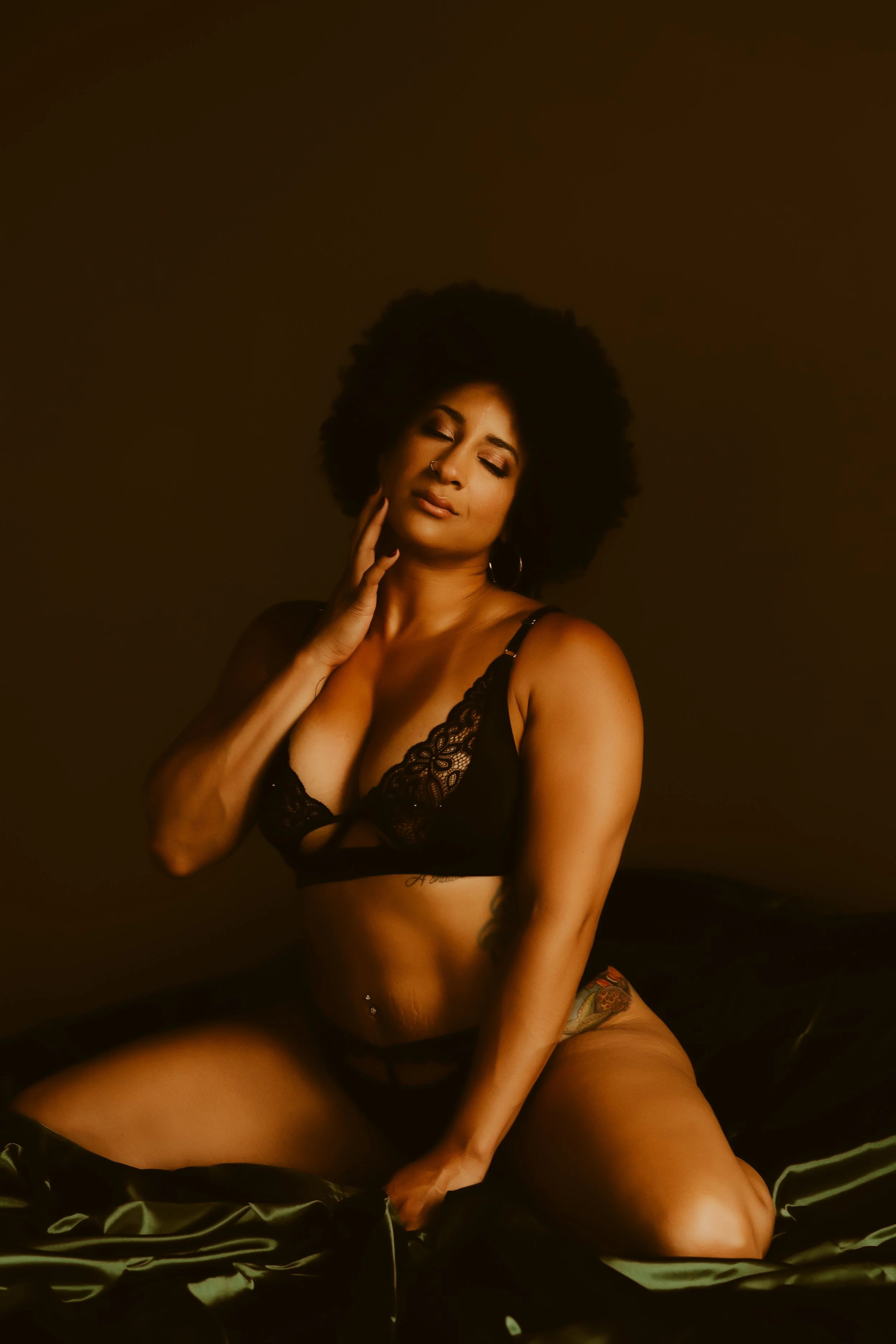 A woman with curly hair wearing black lace lingerie, sitting on a satin sheet with a dark background.