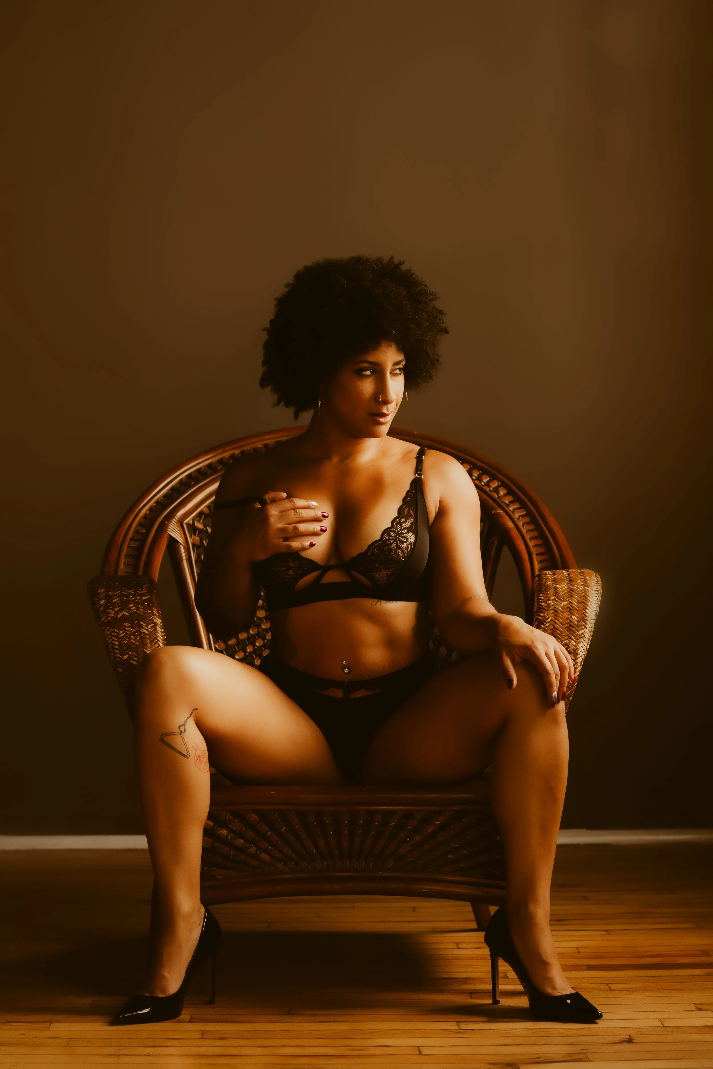 A woman in black lingerie and high heels sitting on a wooden chair against a dark brown background, with one hand on her chest and the other on her thigh.