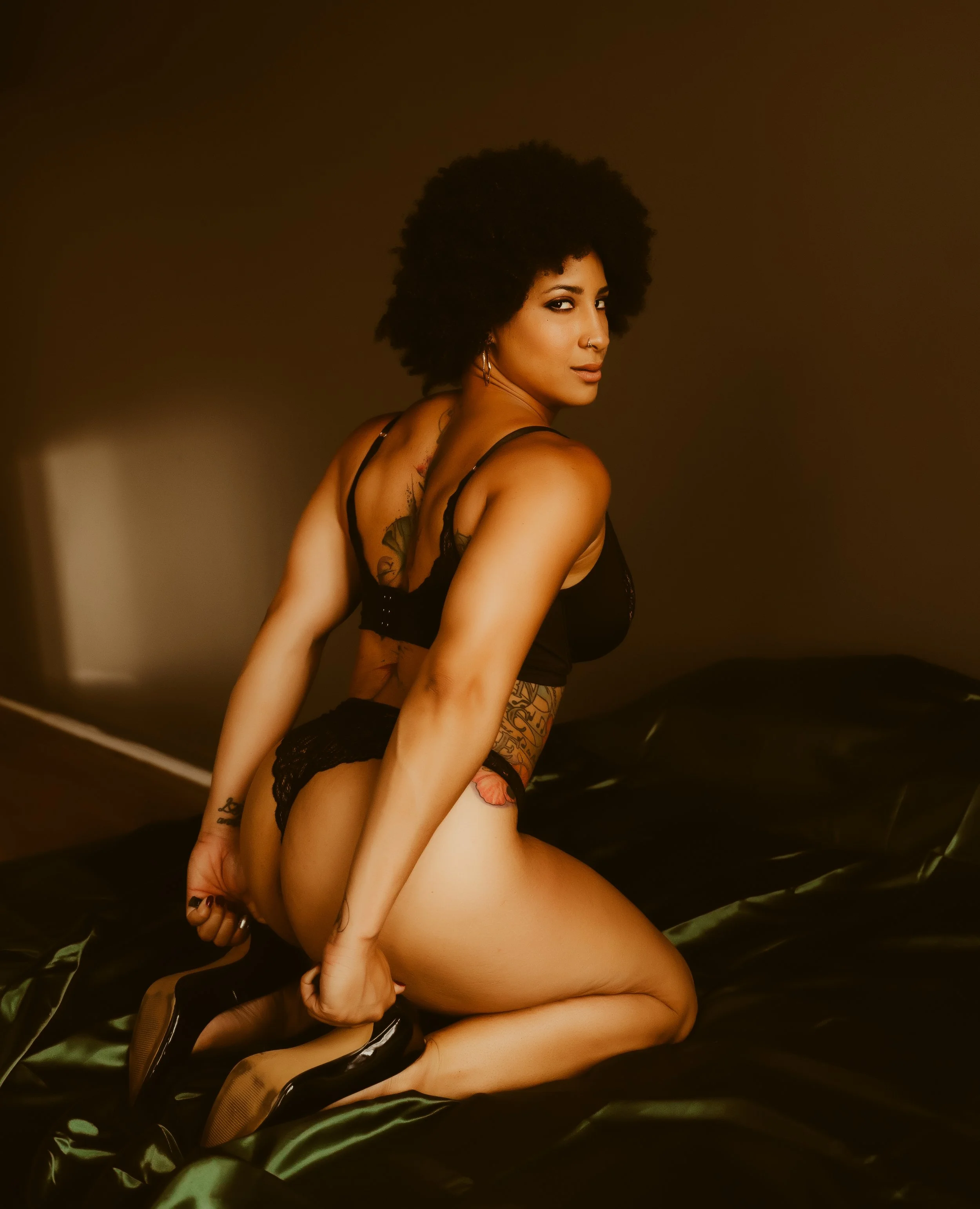 A woman with dark curly hair kneeling on a bed in black lingerie and high heels, looking over her shoulder, with tattoos visible on her back and arm, in a dimly lit room.