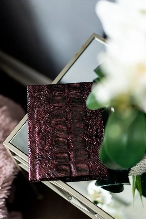 Close-up of a burgundy crocodile-embossed leather wallet placed on a mirror surface with a green vase and white flowers nearby.