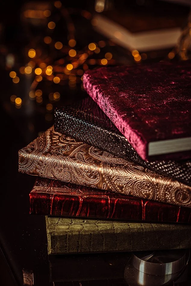 Stack of ornate, decorative books with velvet, embossed, and textured covers in a dimly lit setting with blurred warm string lights in the background.
