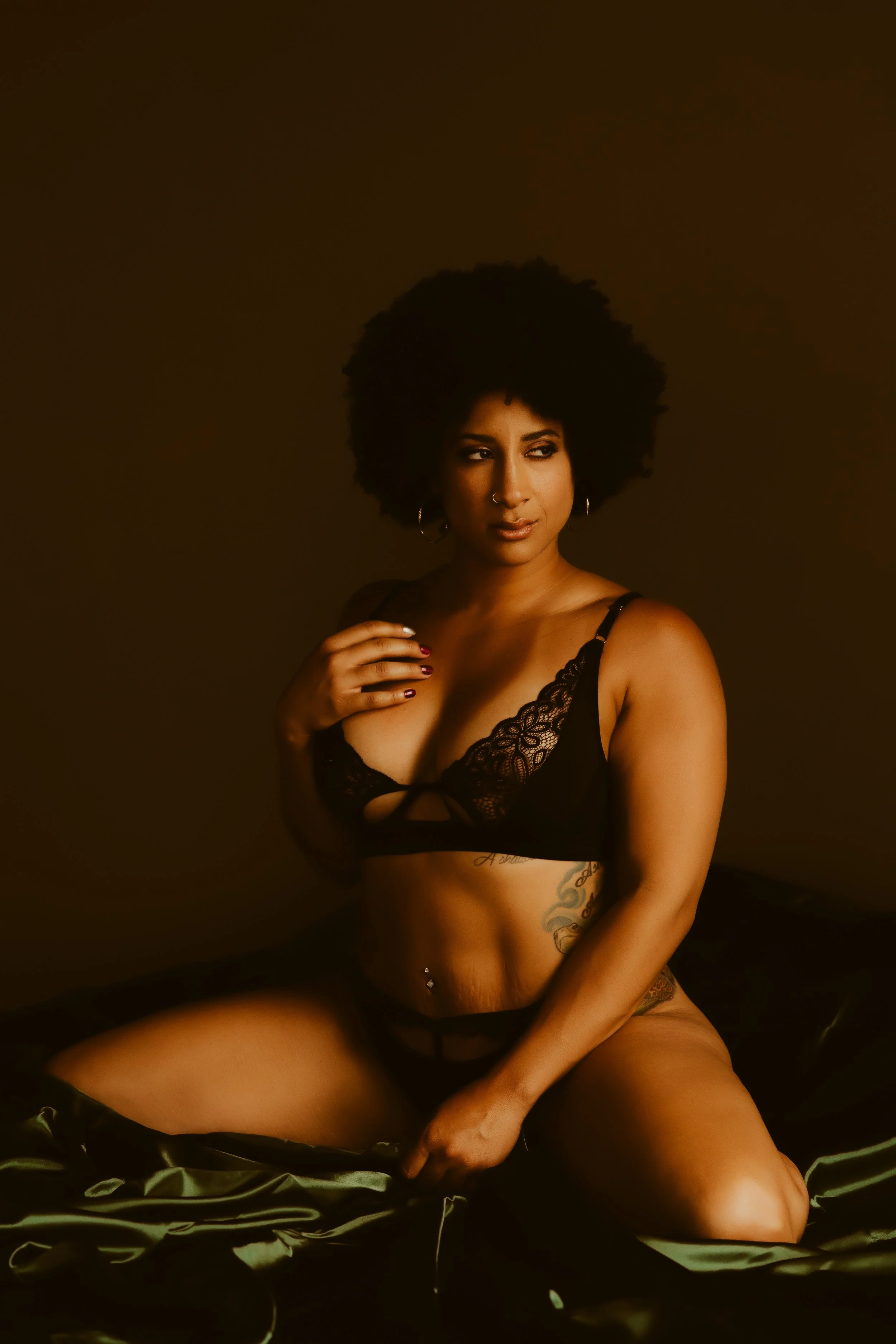 A woman with curly hair wearing black lingerie, kneeling on a bed with black satin sheets, against a dark background.