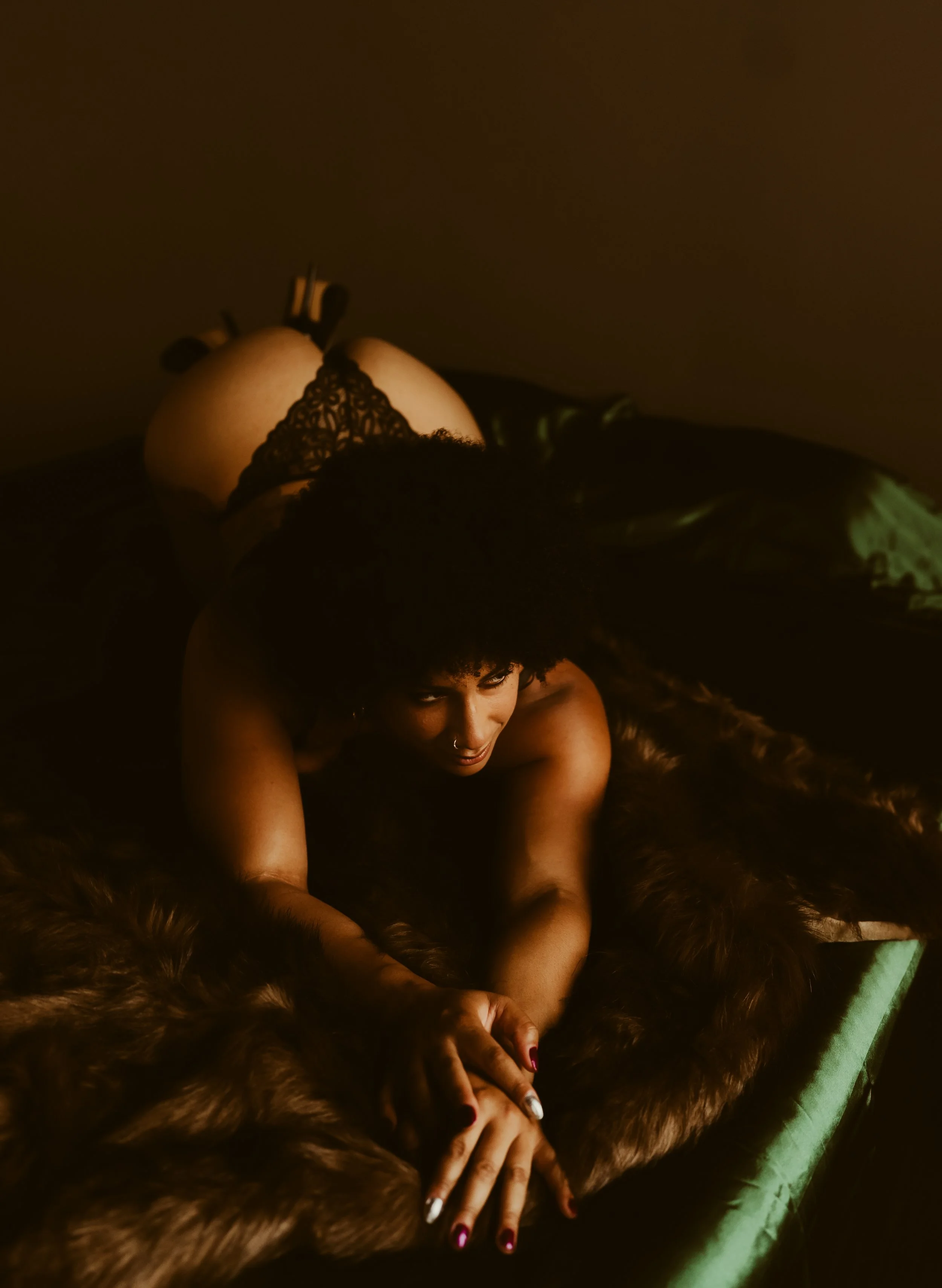 A woman with curly hair lying on her stomach on a bed covered in a fur blanket, looking at the camera.