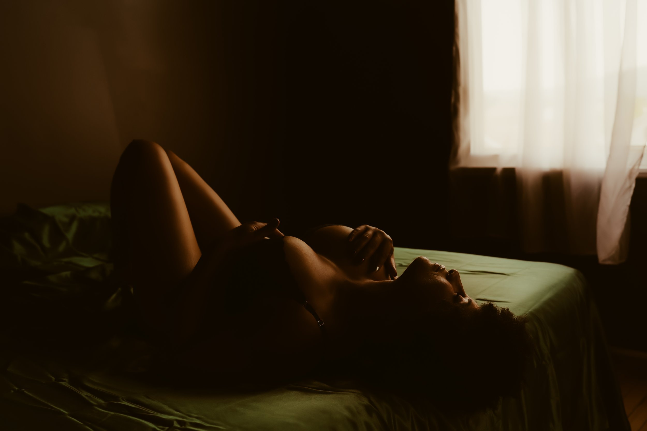 A woman lying on a bed in a dimly lit room, with light coming through a window with curtains.