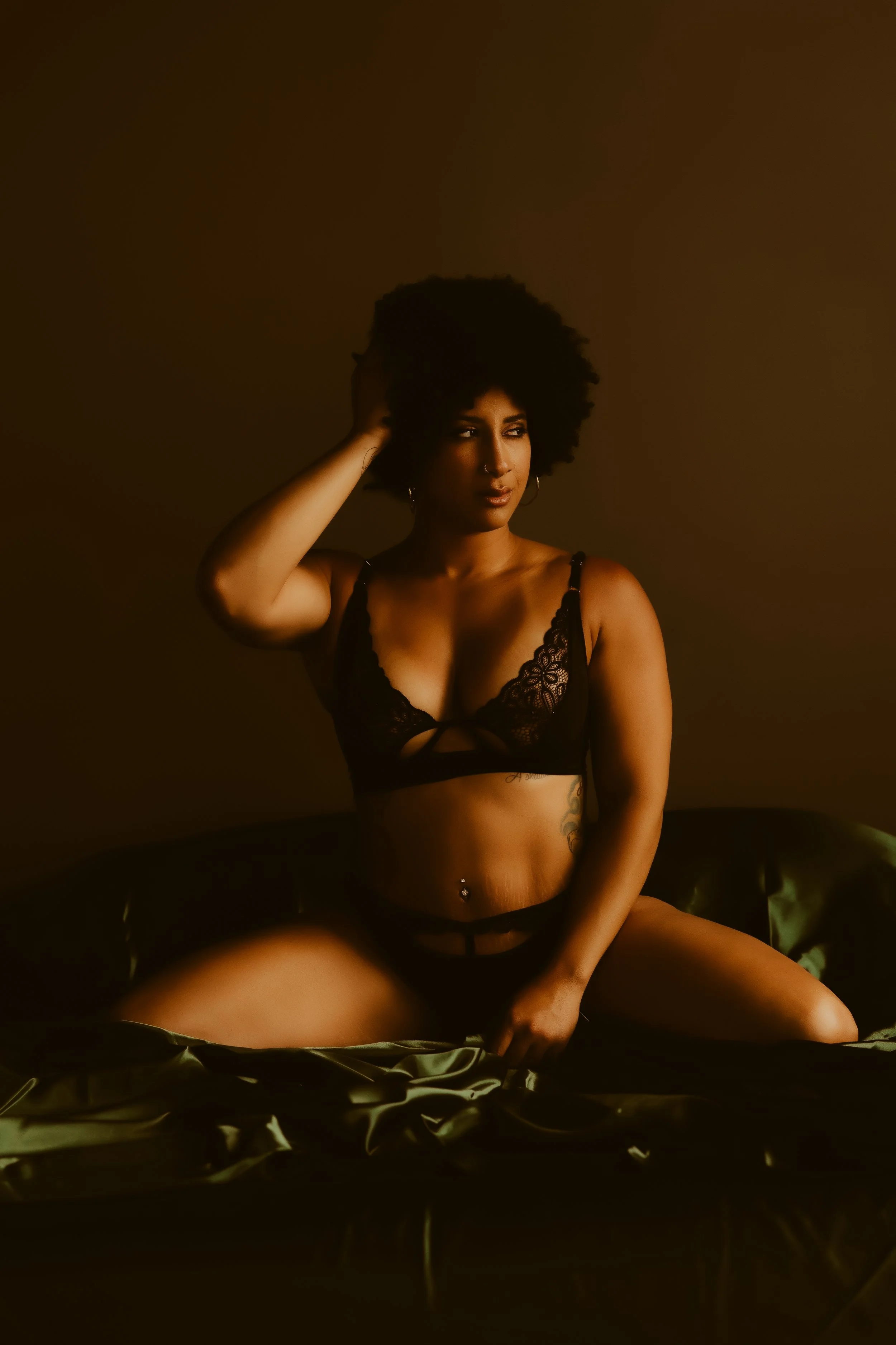 A woman with curly hair sitting on a bed in black lingerie, posing with one hand behind her head.