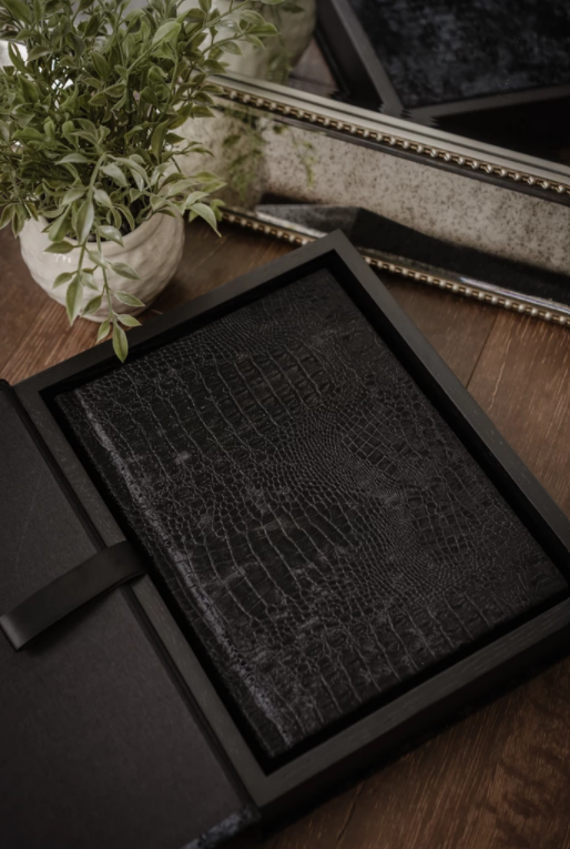 A black leather-bound notebook with a crocodile texture inside a black presentation box, placed on a wooden surface next to a small green plant and a mirror.