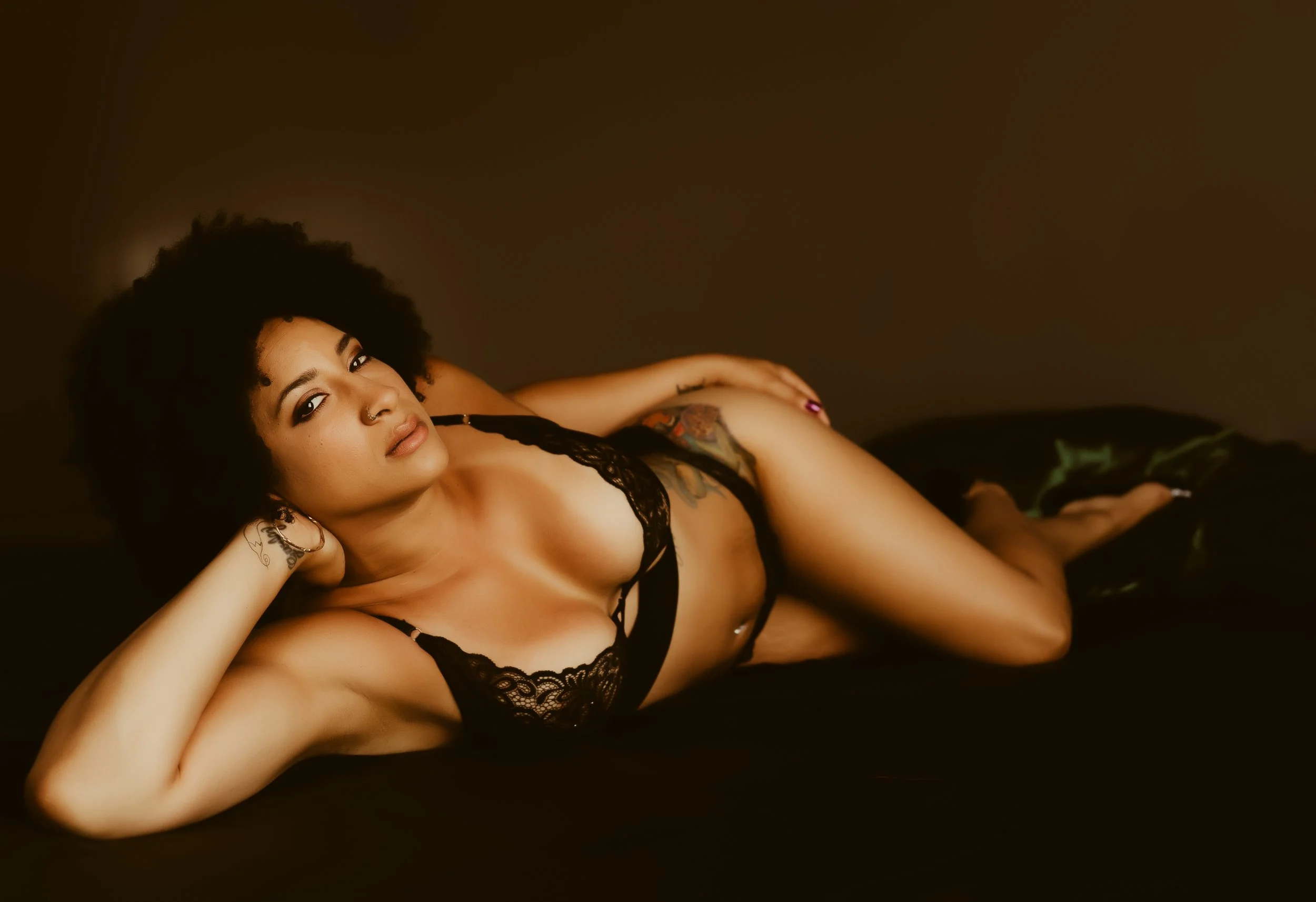 A woman with curly hair lying on a dark surface, wearing black lace lingerie, with tattoos visible on her arm and torso, looking at the camera.
