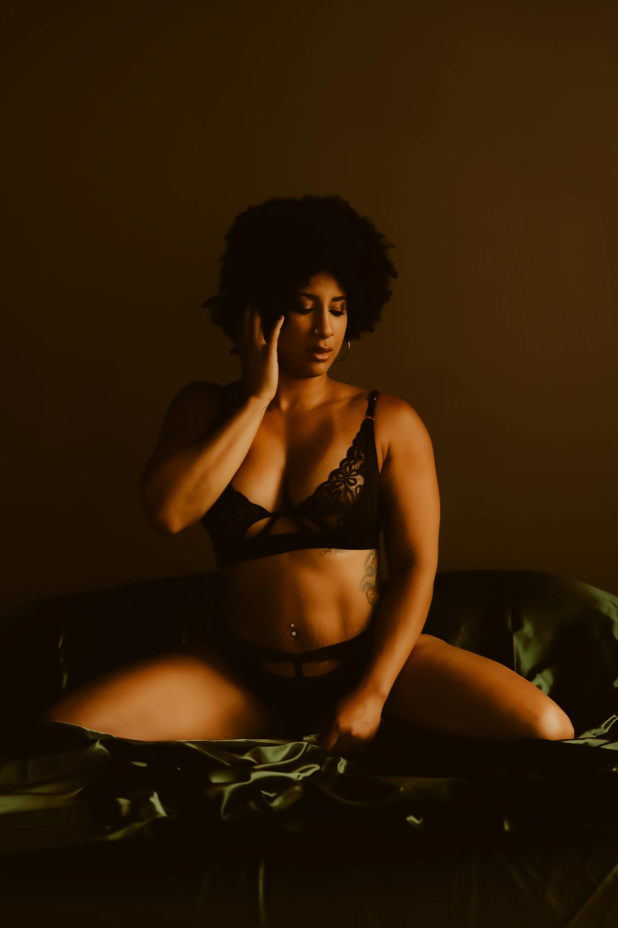 A woman with afro hair sitting on a bed in black lace lingerie, with a dark background and satin sheets.
