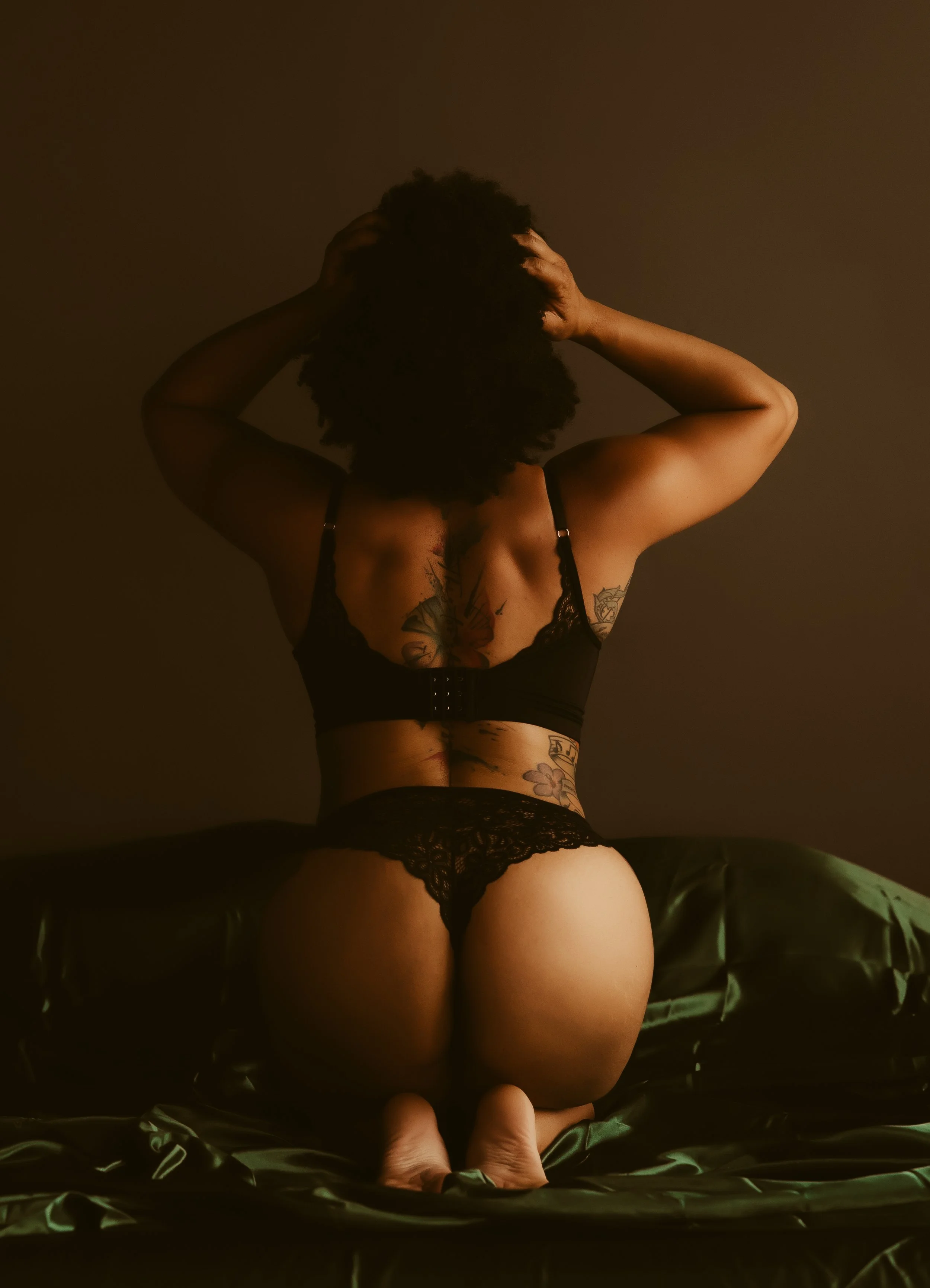 A person with curly hair kneeling on a bed with black satin sheets, facing away from the camera, wearing black lace lingerie, with tattoos on their back and side.