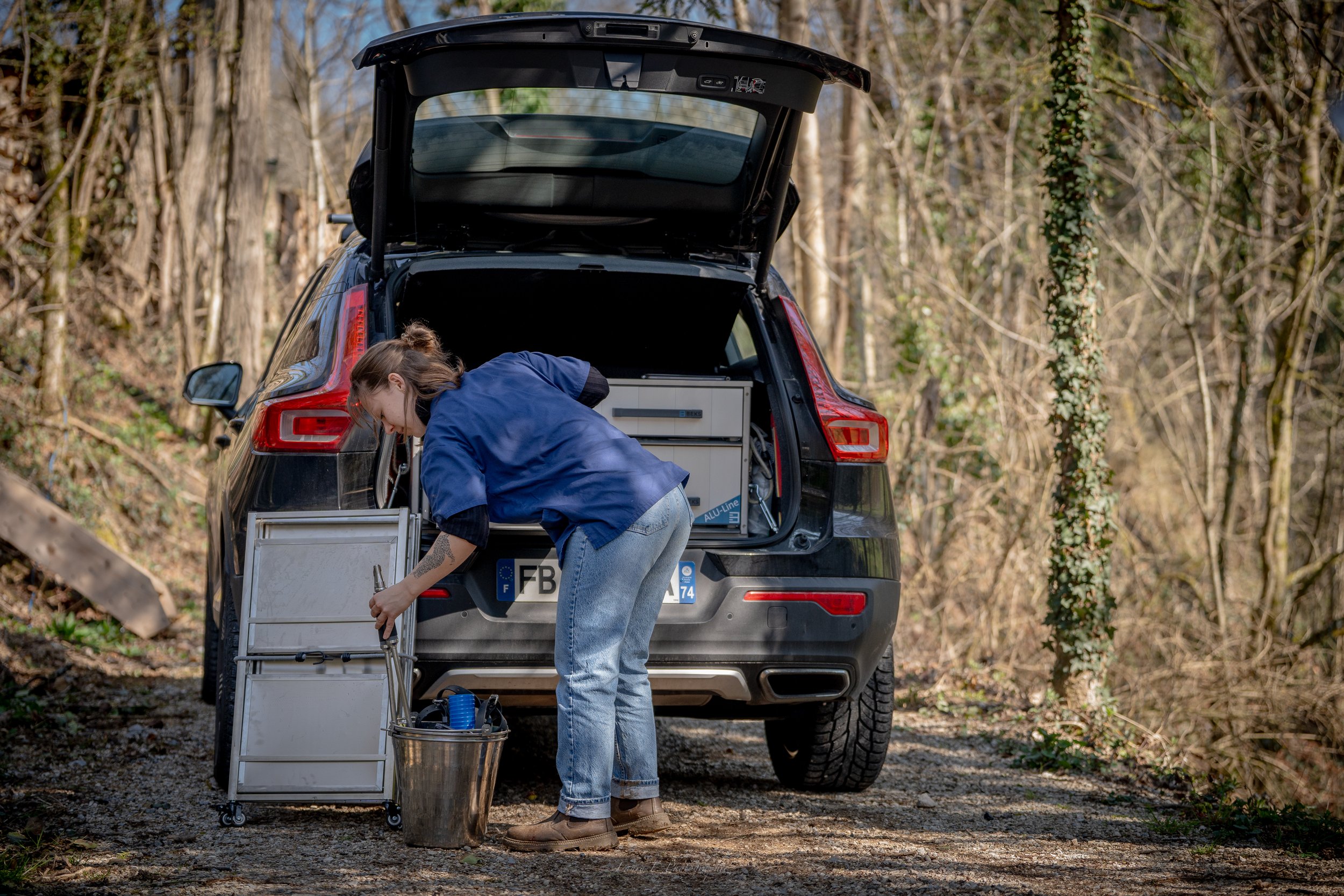 Une femme effectue une réparation en plein air à l'arrière d'une voiture noire dans une forêt. La femme porte une veste bleue et des jeans.