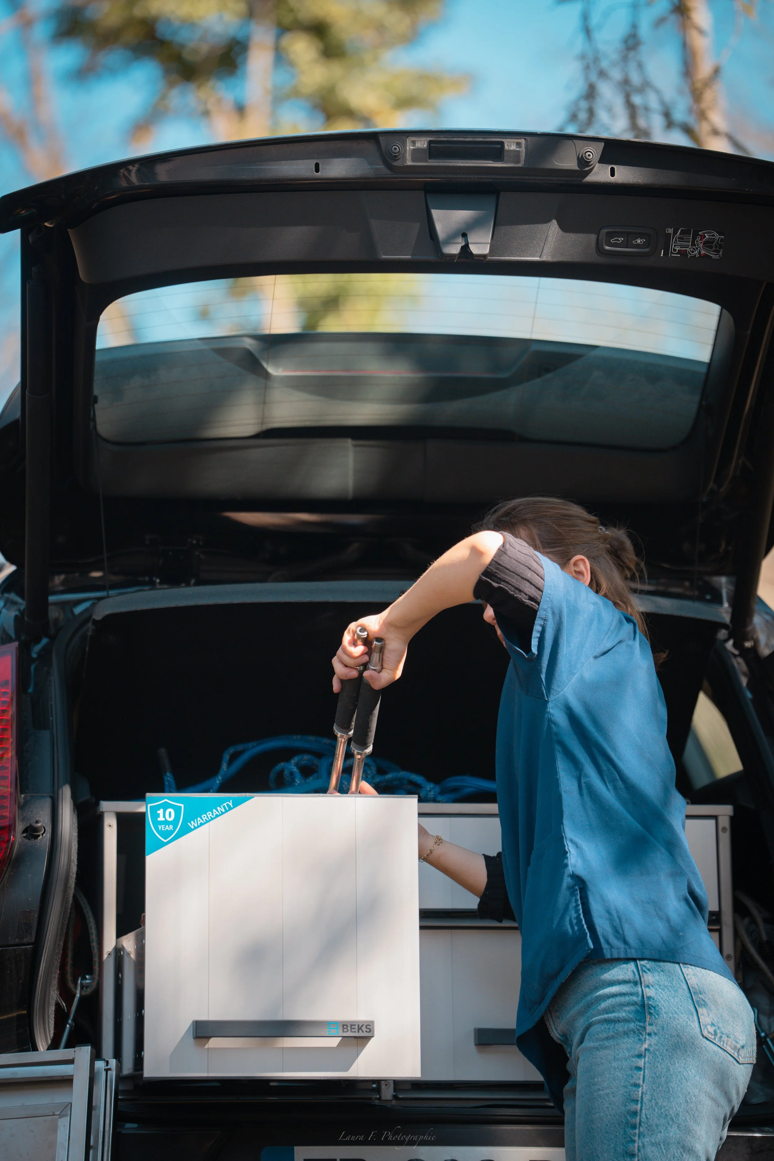 Une femme effectue des réparations sur une voiture électrique avec un coffre ouvert, utilisant des outils pour manipuler la batterie.