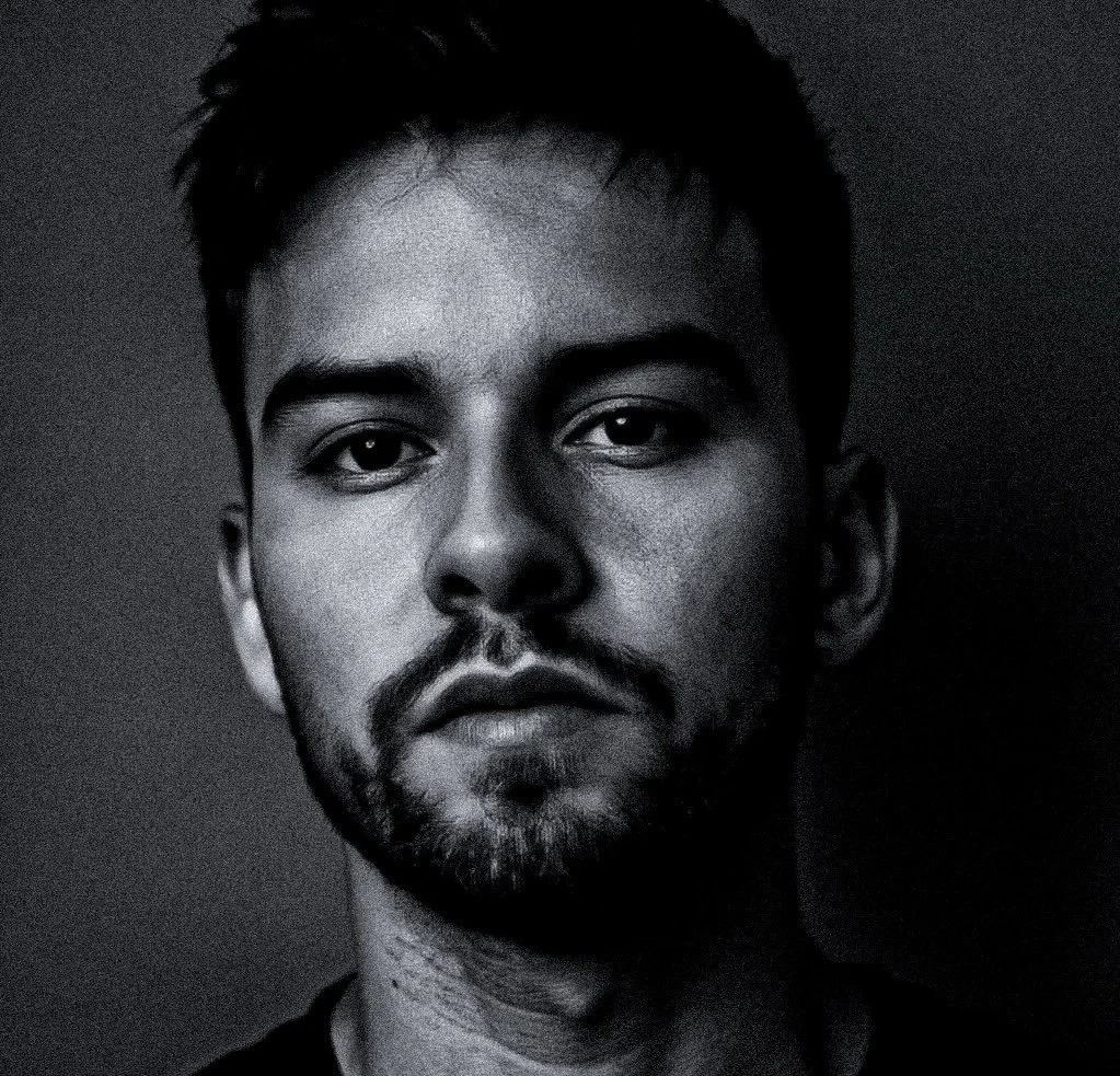 Close-up black and white portrait of a young man with dark hair and a beard, looking directly at the camera with a serious expression.