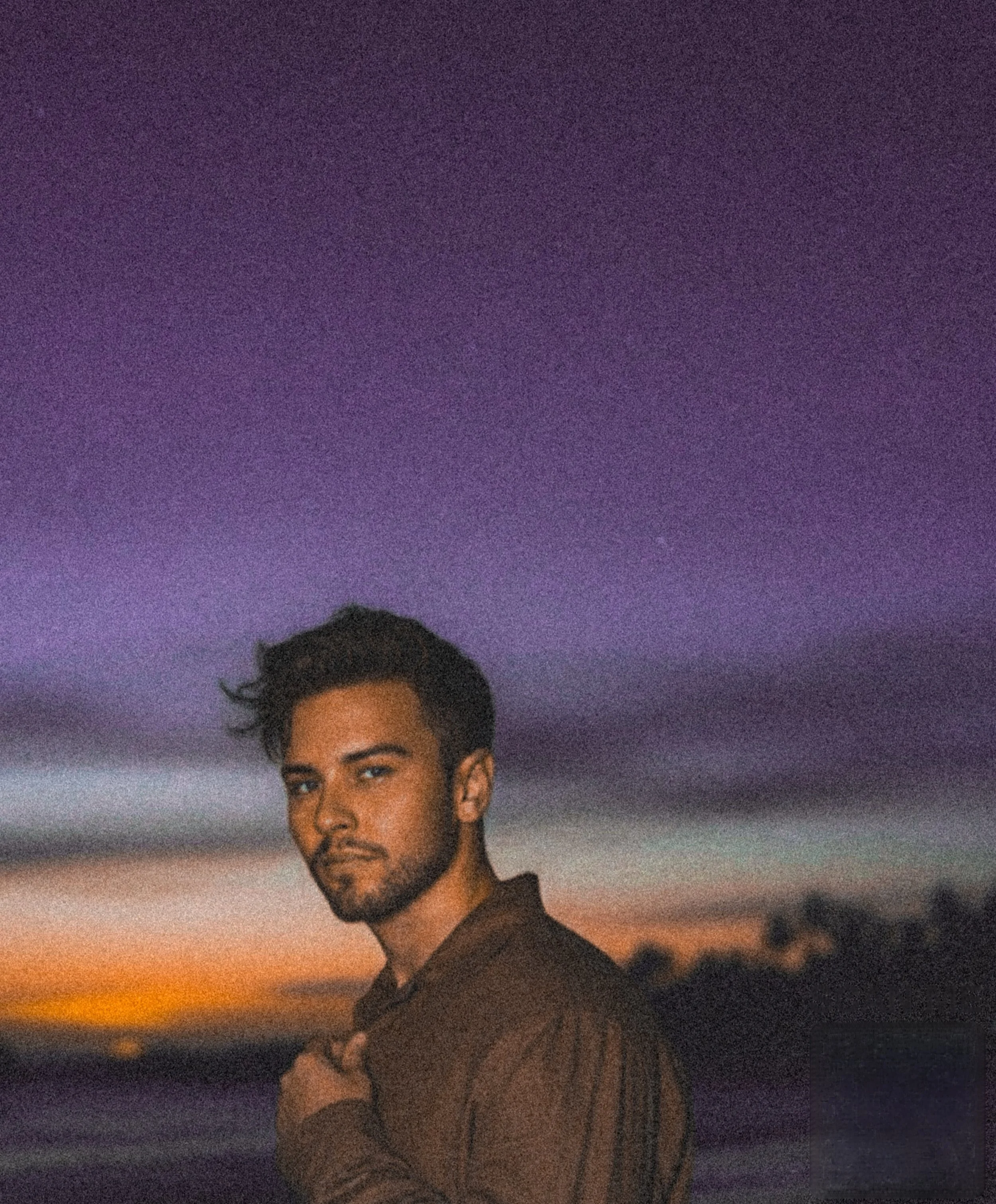 A young man with dark hair and beard standing outdoors during sunset, with a colorful sky of purple, pink, and orange hues in the background.