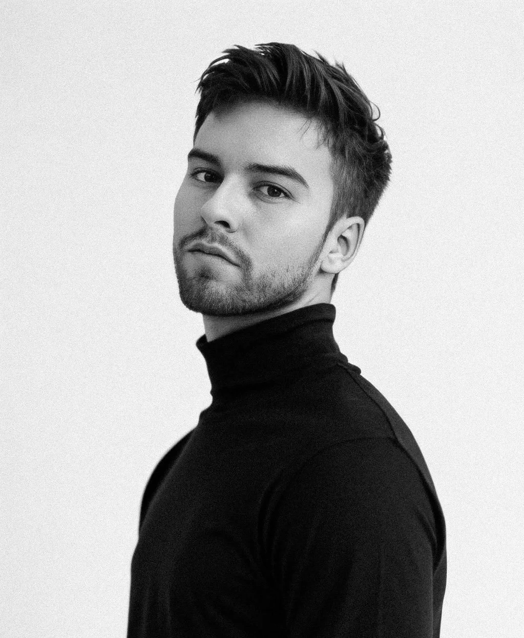 Black and white portrait of a young man with short styled hair and light stubble, wearing a black turtleneck, looking at the camera with a slight tilt of the head, against a plain light background.