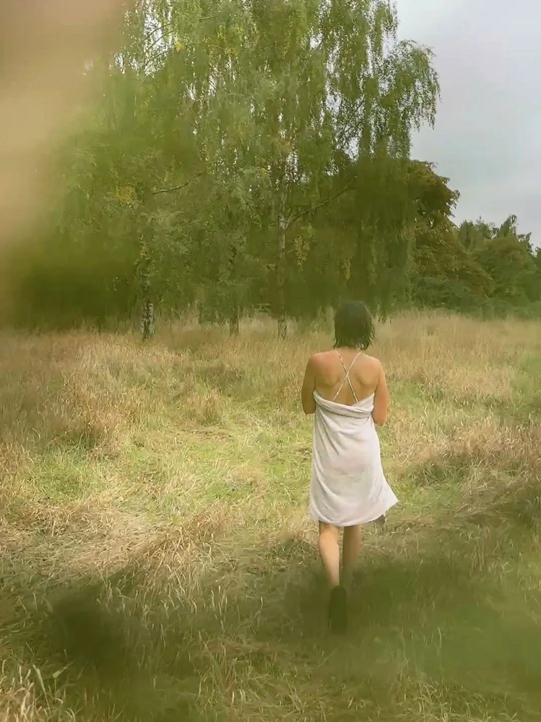 A person with black hair is walking away in a field of tall grass, wearing a white dress and surrounded by trees under a cloudy sky.