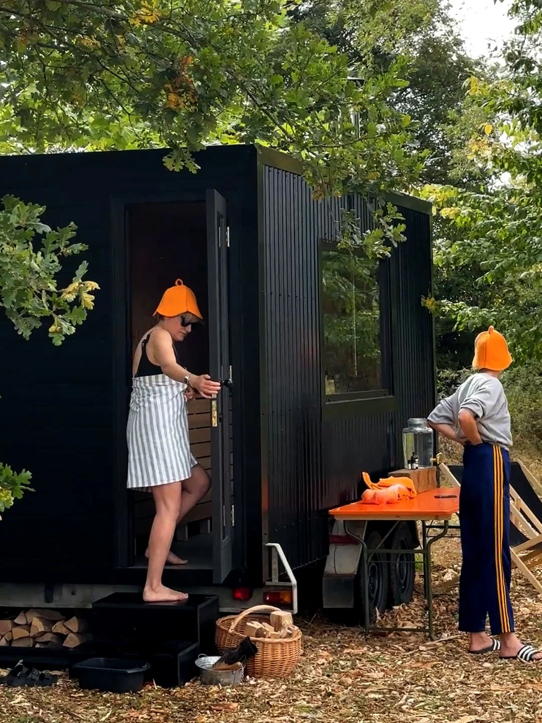 Two people outside a surrounded by trees. The woman is coming out of the cabin wearing a striped apron dress, sunglasses, and an orange hat shaped like a bell. The person outside, wearing a gray hoodie, blue pants with yellow stripes, striped slipper
