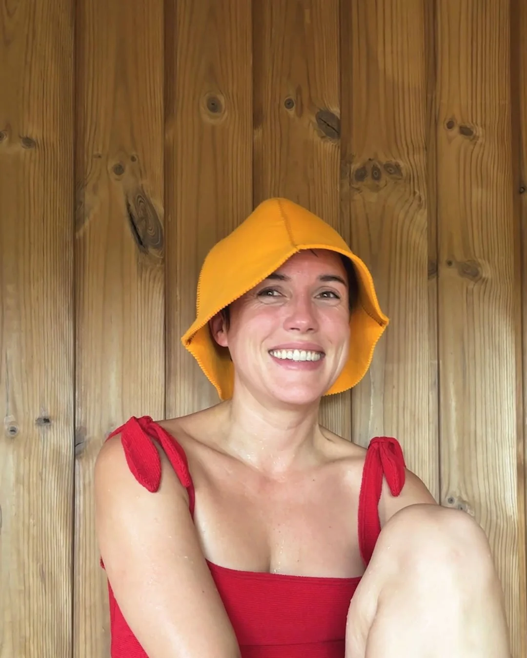 A woman smiling, wearing a yellow hat and a red tank top, sitting against a wooden wall.