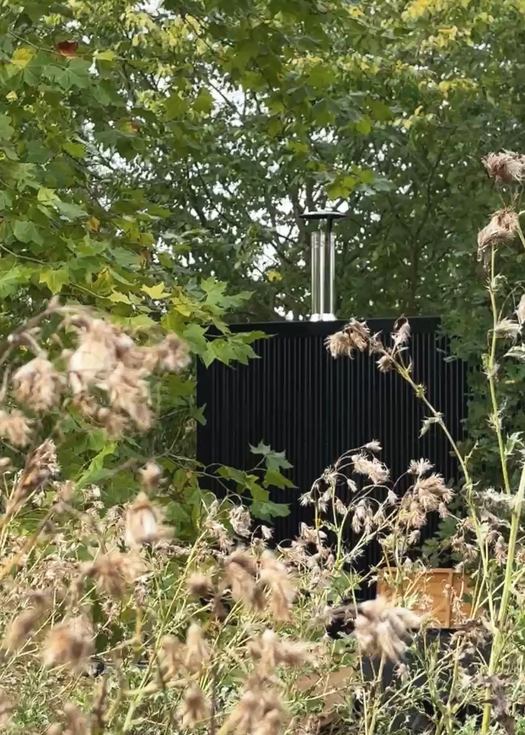 Black wood-fired mobile sauna surrounded by plants and green trees.