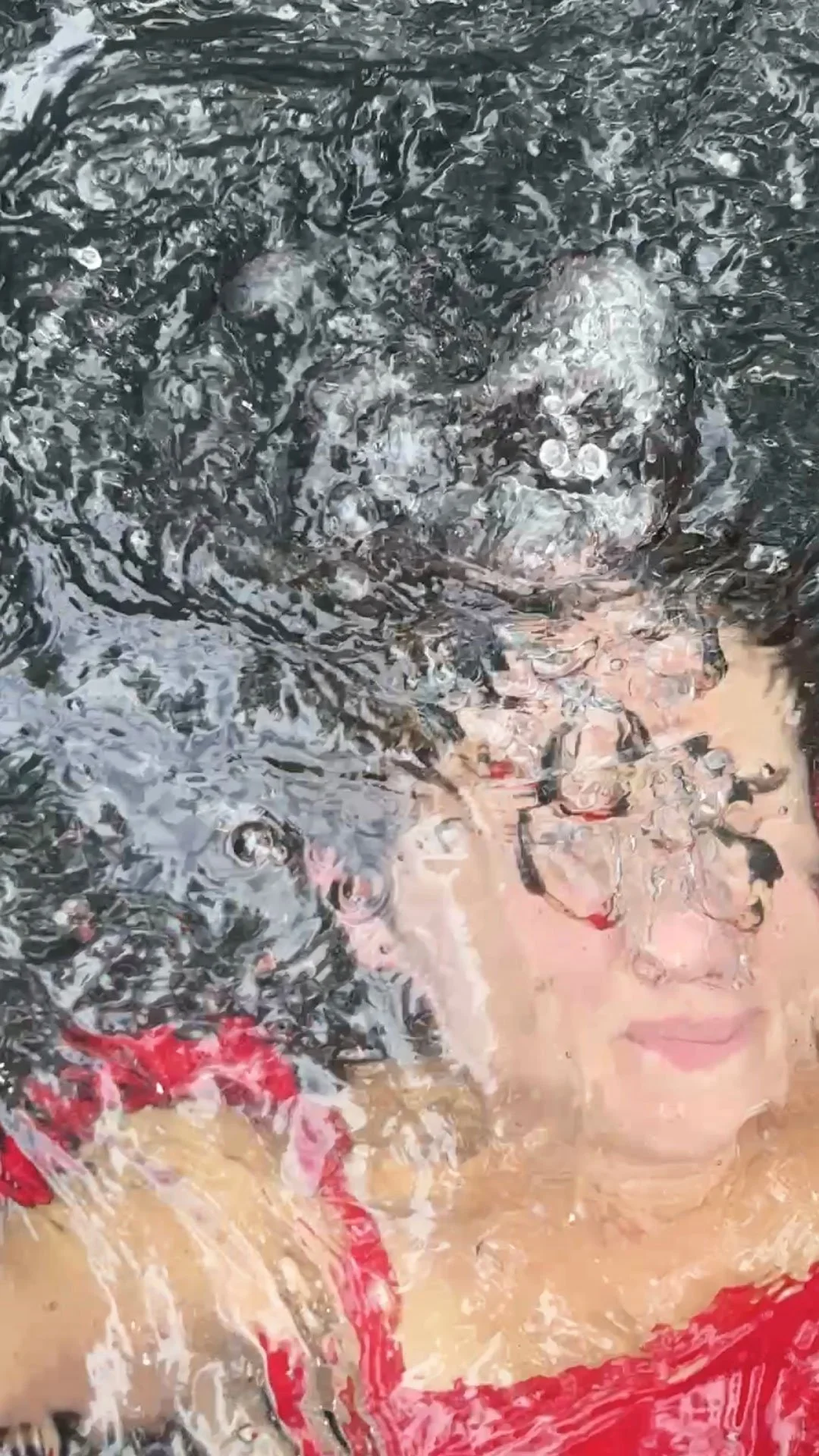 Person lying underwater with their face and body partially visible through rippling water, wearing a red top.