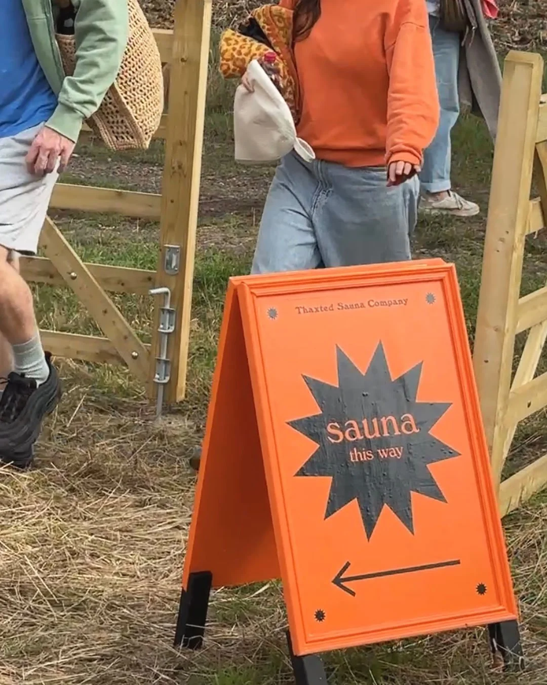 An orange sign with black text and an arrow pointing left, indicating the way to the sauna at an outdoor location. People are walking around, and there's a wooden fence or gate in the background.