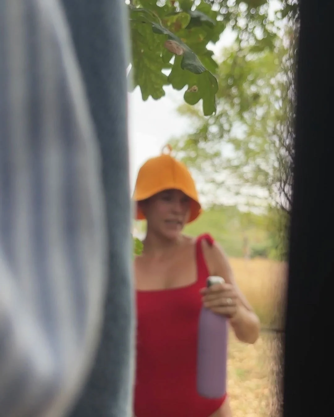 A woman wearing an orange sauna hat and a red tank top, holding a purple water bottle and looking at her phone outdoors, surrounded by green foliage and trees.