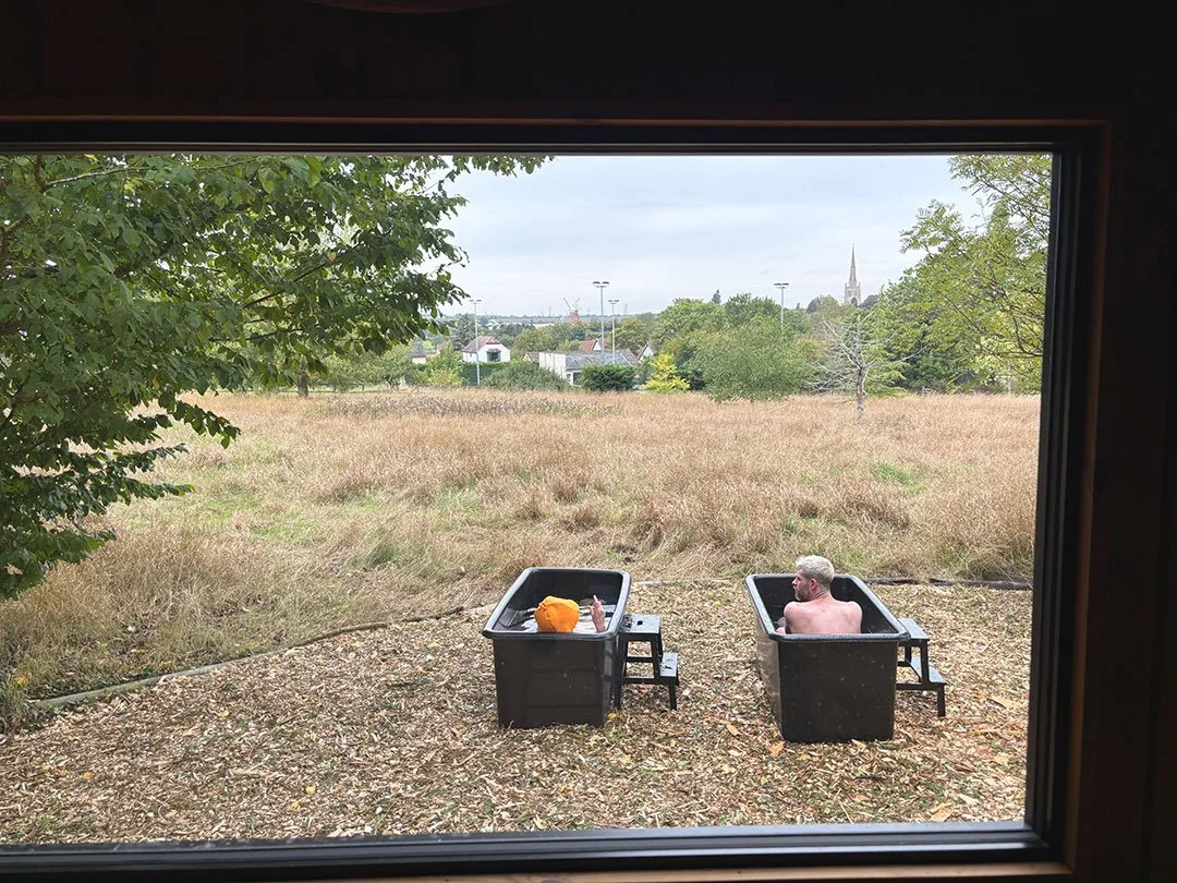 Two people sitting in black outdoor tubs with water, viewed through a window; one person has a gray beard, and the other person is a child with an orange hat. The tubs are on a dirt ground overlooking a field with trees and a neighborhood in the back