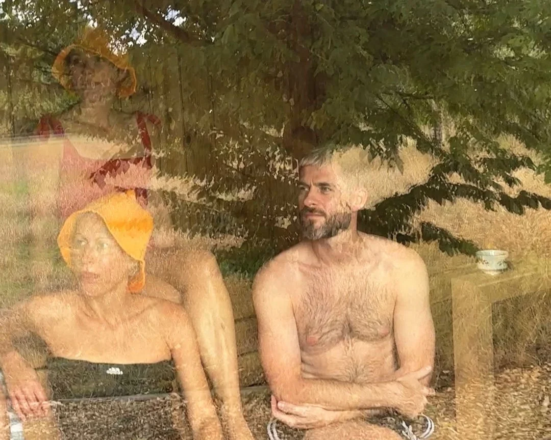 A reflection of three people, two women and one man, seen through a glass window with a tree behind them. All have wet hair and appear to be sitting outdoors.