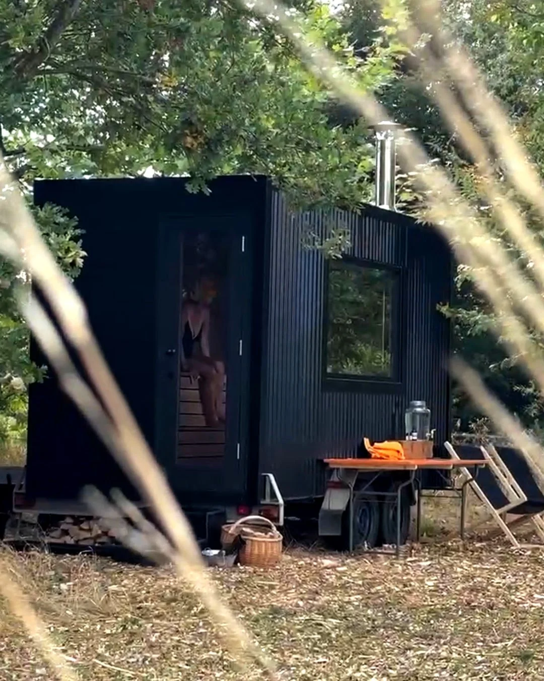 A black wood-fired sauna on wheels set in a wooded area, with a small outdoor table holding a jar and a cloth, a basket on the ground, and a woman sitting inside, partially visible through the door.