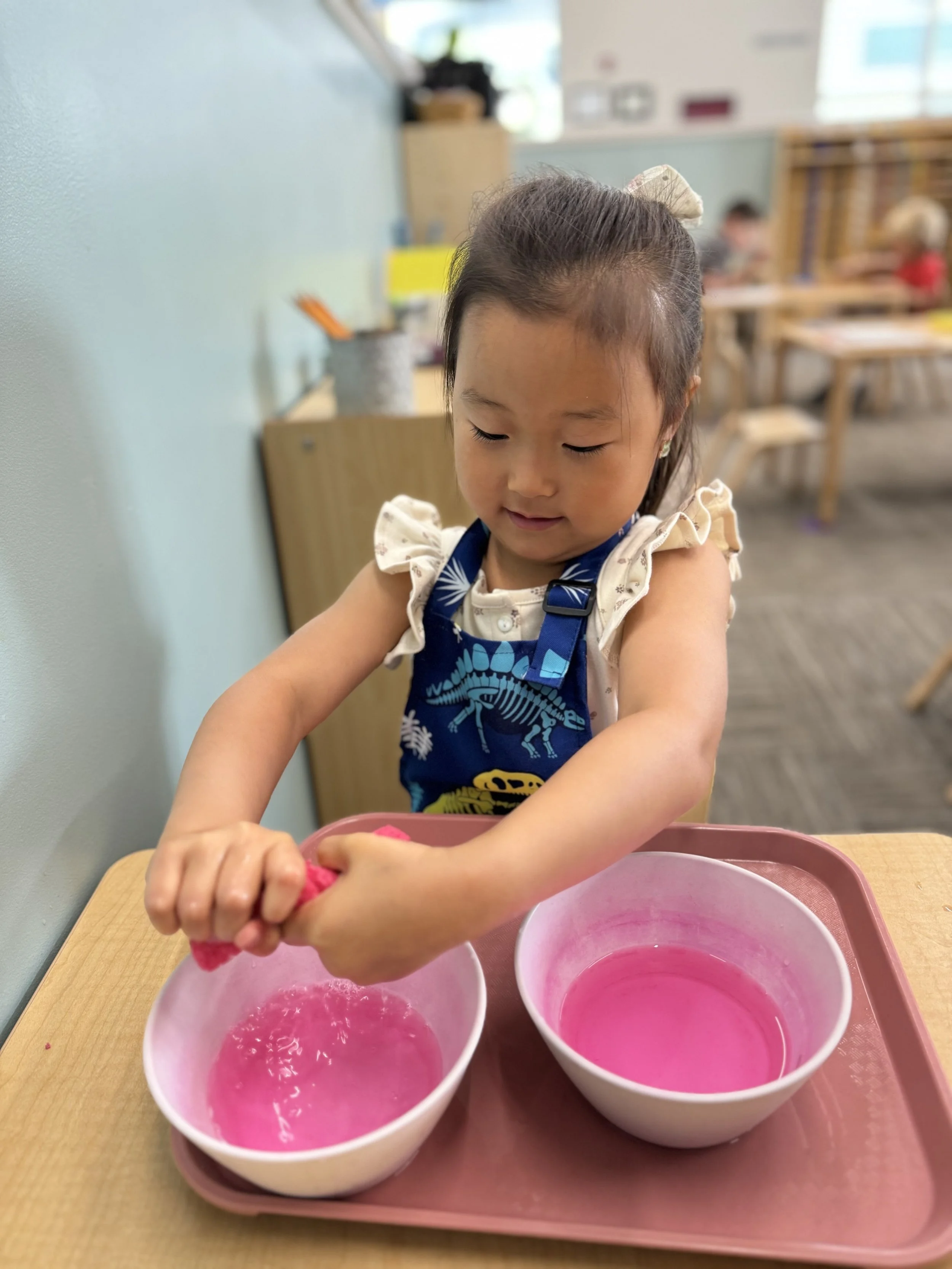 A young girl wearing a dark blue apron with dinosaur prints is kneading pink slime in a white bowl, sitting at a wooden table in a classroom setting.