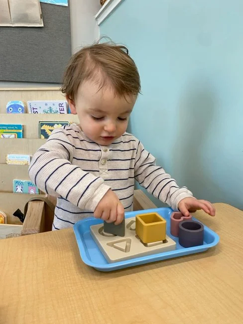 A young child playing with shape-sorting toys on a wooden table in a classroom or play area.