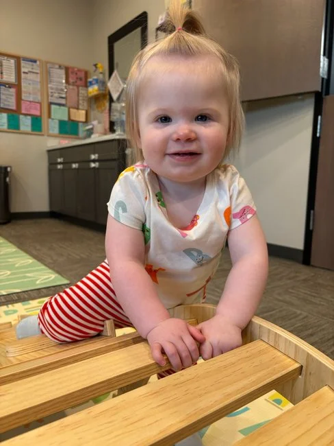 A young girl with blonde hair tied in a ponytail, wearing a colorful animal print shirt and red striped pants, is climbing on a wooden play structure indoors, smiling at the camera.