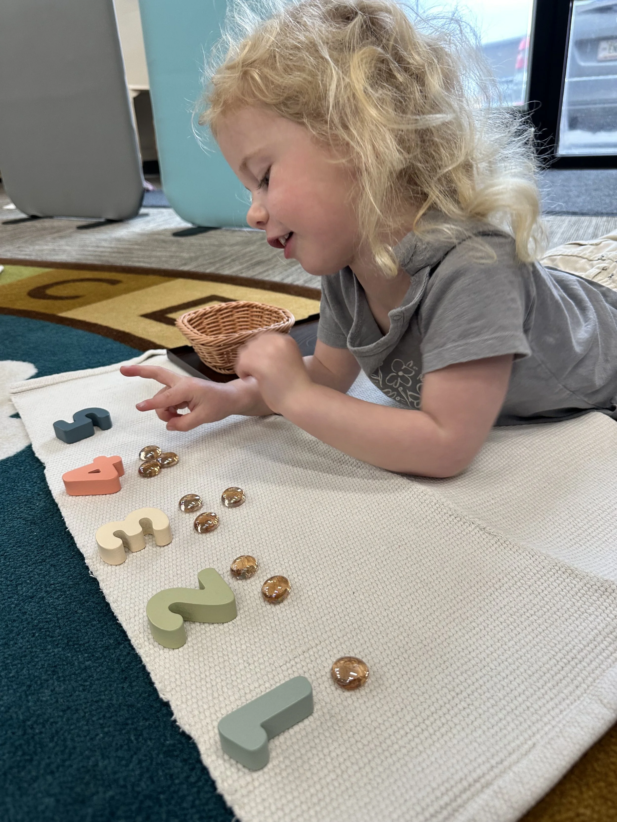 A young girl with curly blonde hair lying on her stomach on a woven mat, arranging colorful magnetic numbers and glass beads with a smile.