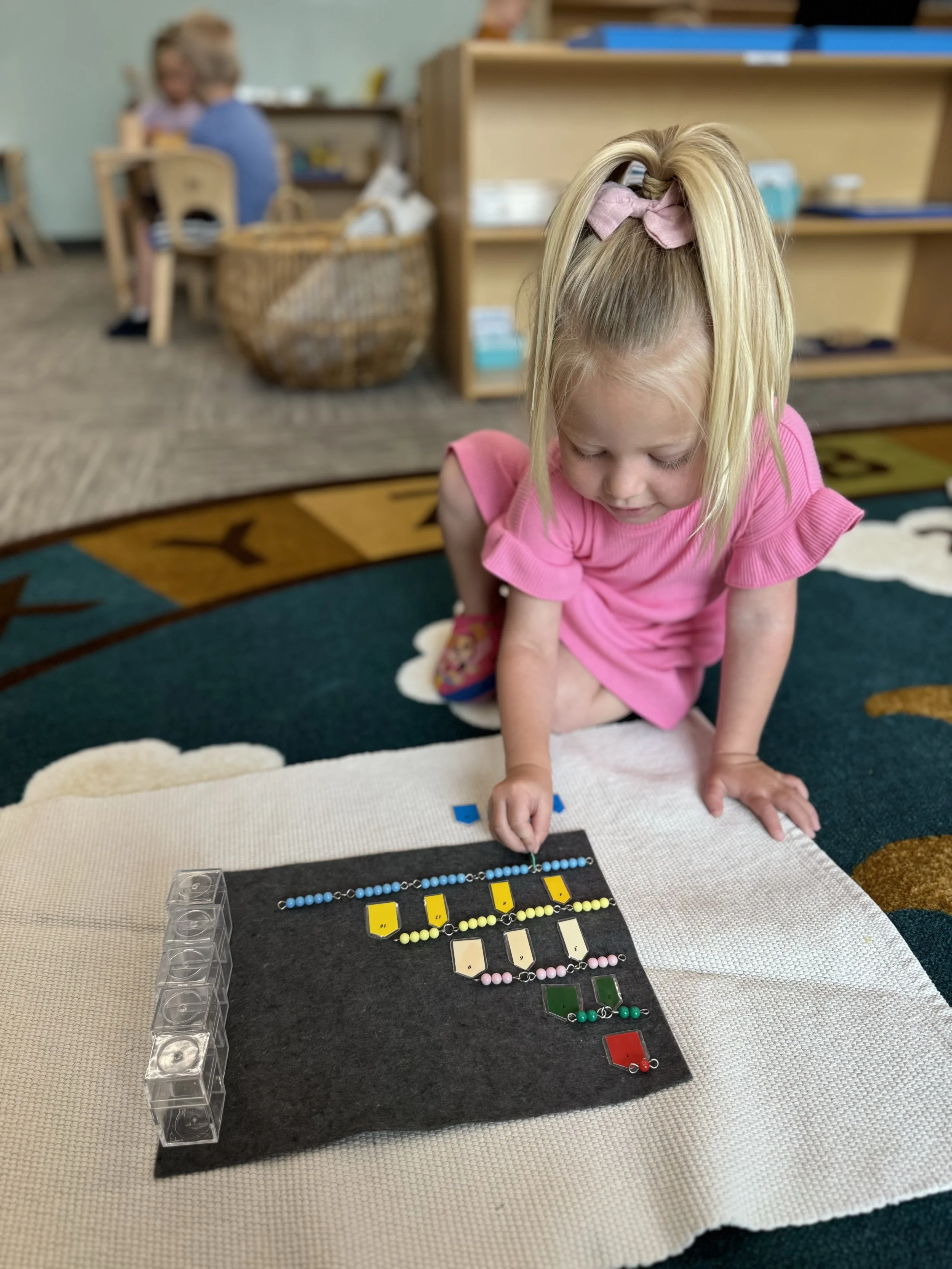 A young girl in a pink dress with a pink bow in her hair, kneeling on the floor, working with a bead sorting activity on a black mat, with colorful beads and key tags, in a classroom setting.