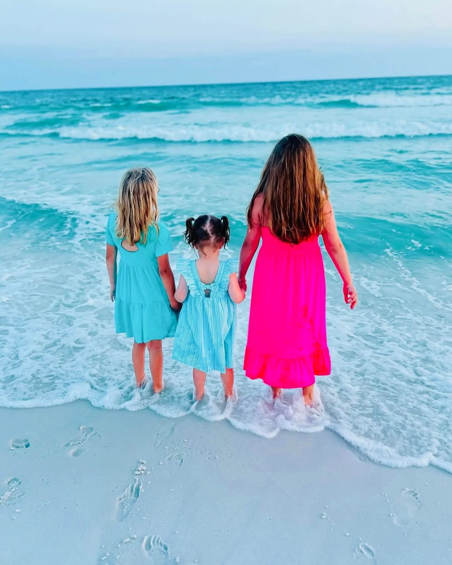 Three girls in colorful dresses holding hands and standing at the shoreline, facing the ocean.