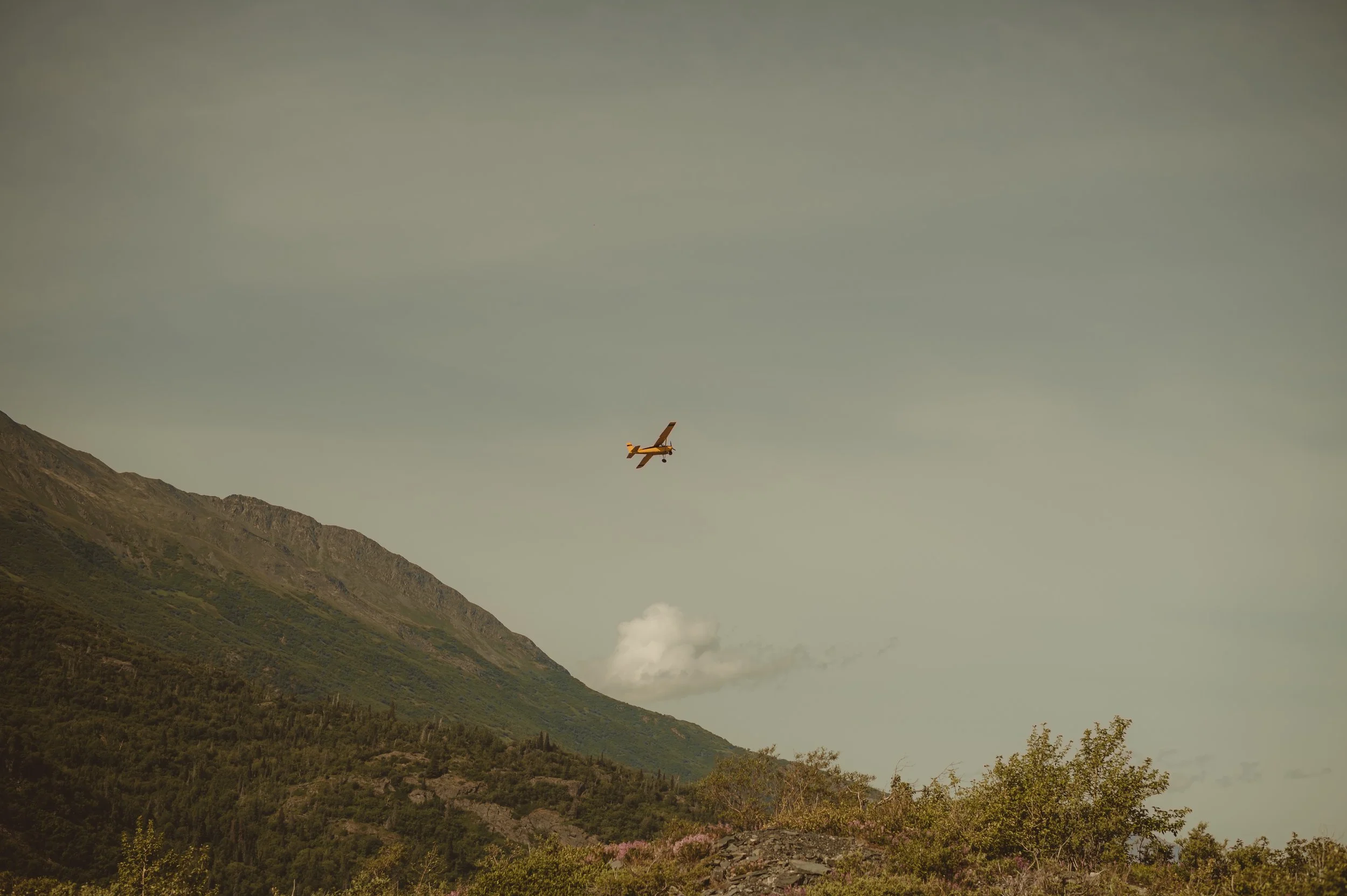 A small yellow and white airplane flying over green mountains.