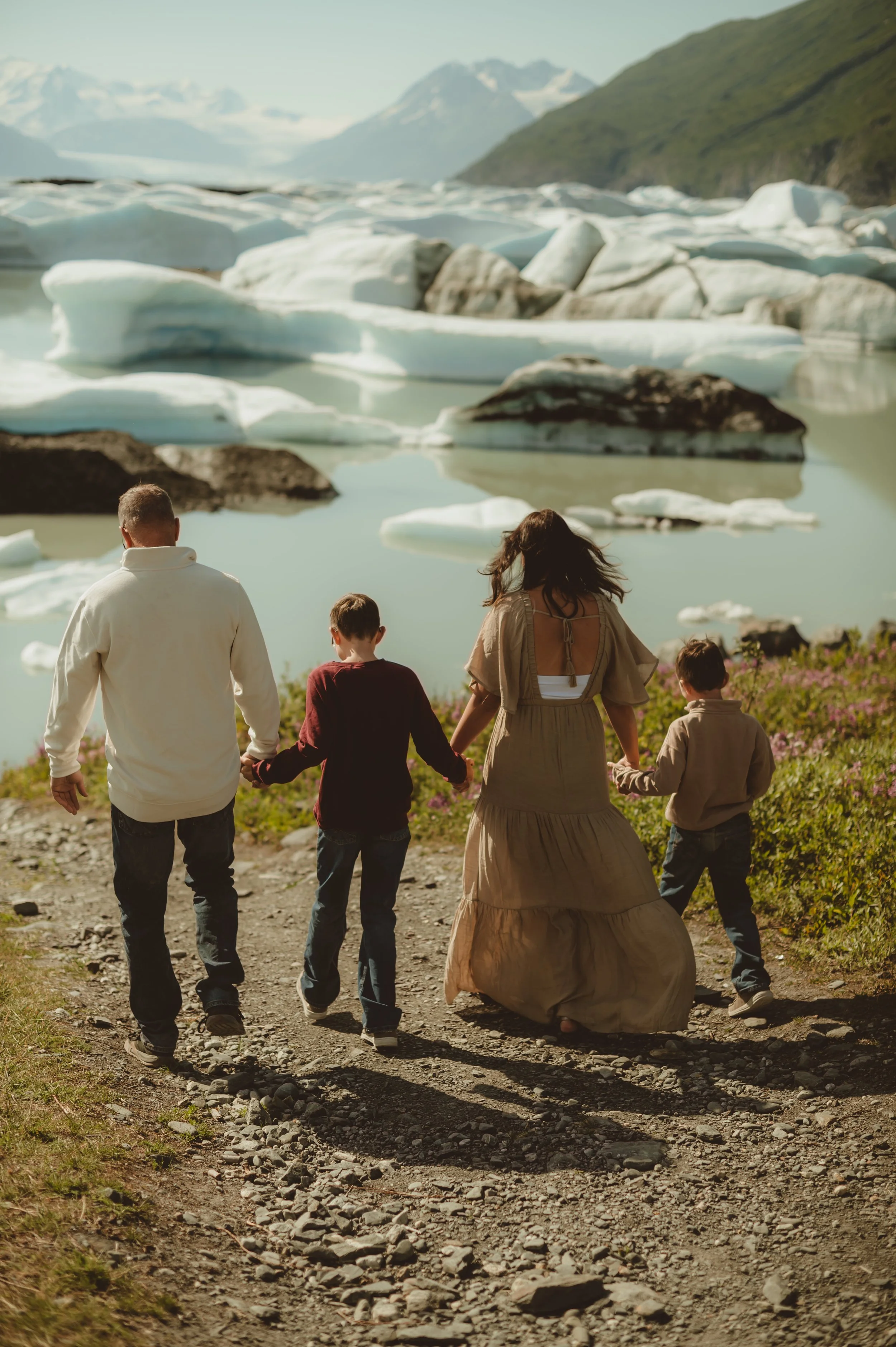 Family walking along a rocky path near a glacial lake with icebergs and mountains in the background.